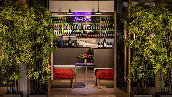 Una zona de bar con estanterías llenas de botellas, dos sillones tapizados de rojo y una mesa con un florero.