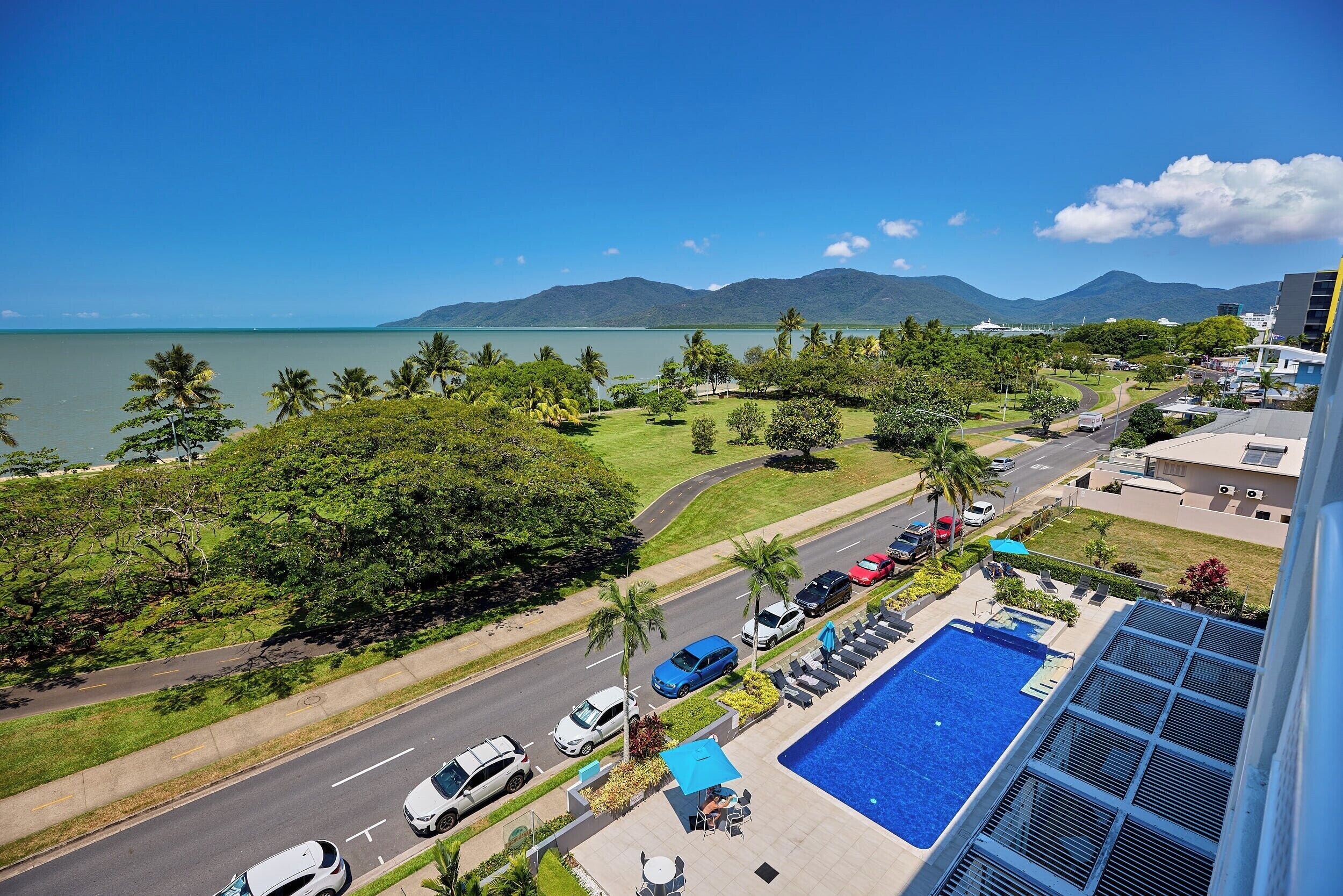 A hotel with a pool, a view of the sea, and a road with parked cars.