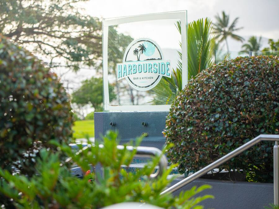 Naamplaatje voor 'Harbourside Bar & Kitchen' met een palmboomlogo, omringd door begroeiing en een metalen hekwerk.