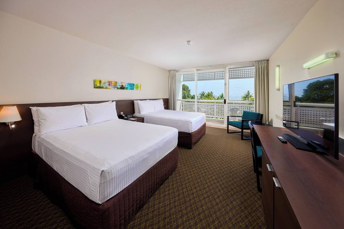 Habitación de hotel con dos camas, un escritorio, un televisor y un balcón con vistas a palmeras.