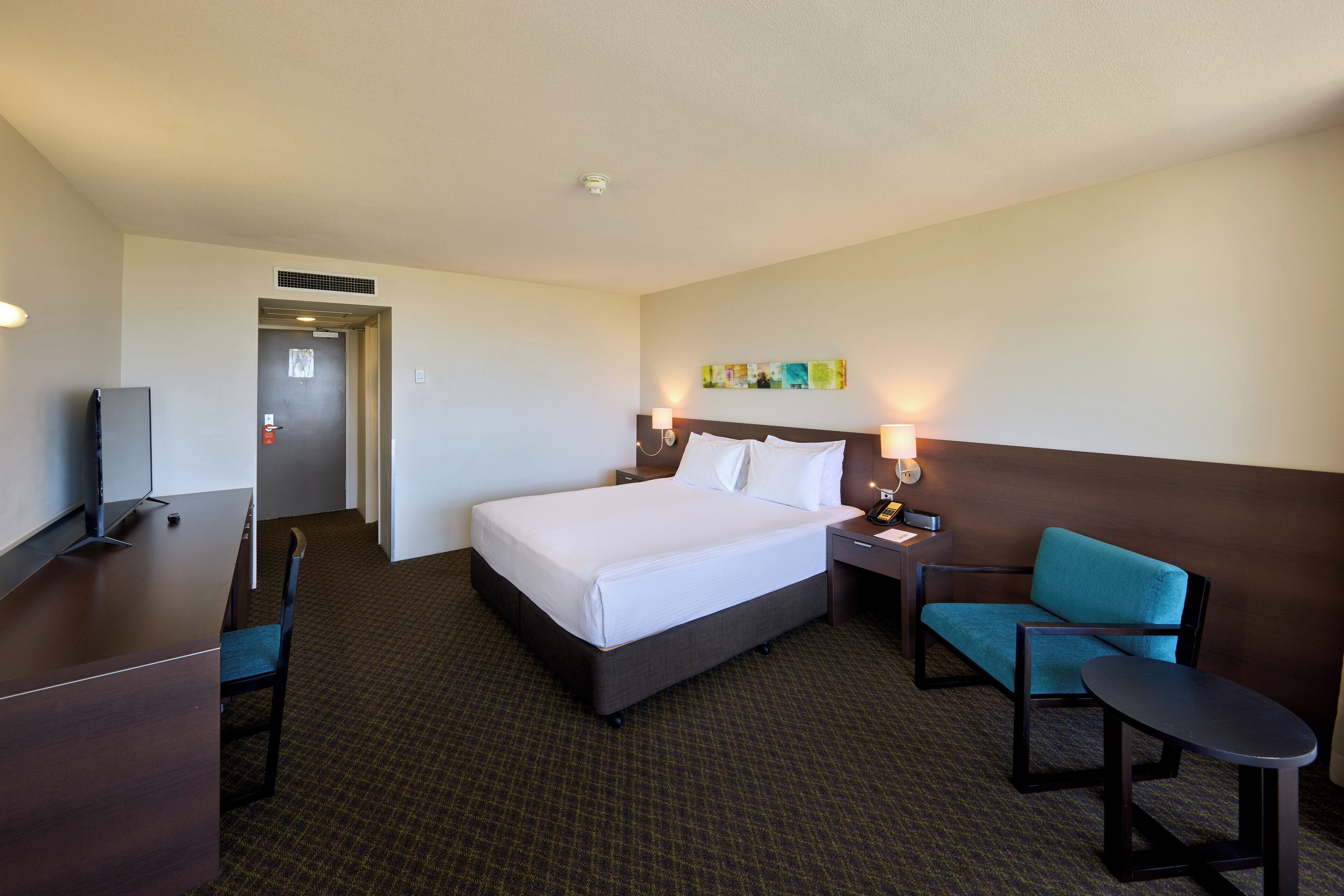 A hotel room with a bed, a desk, a chair, a small table, and a TV.