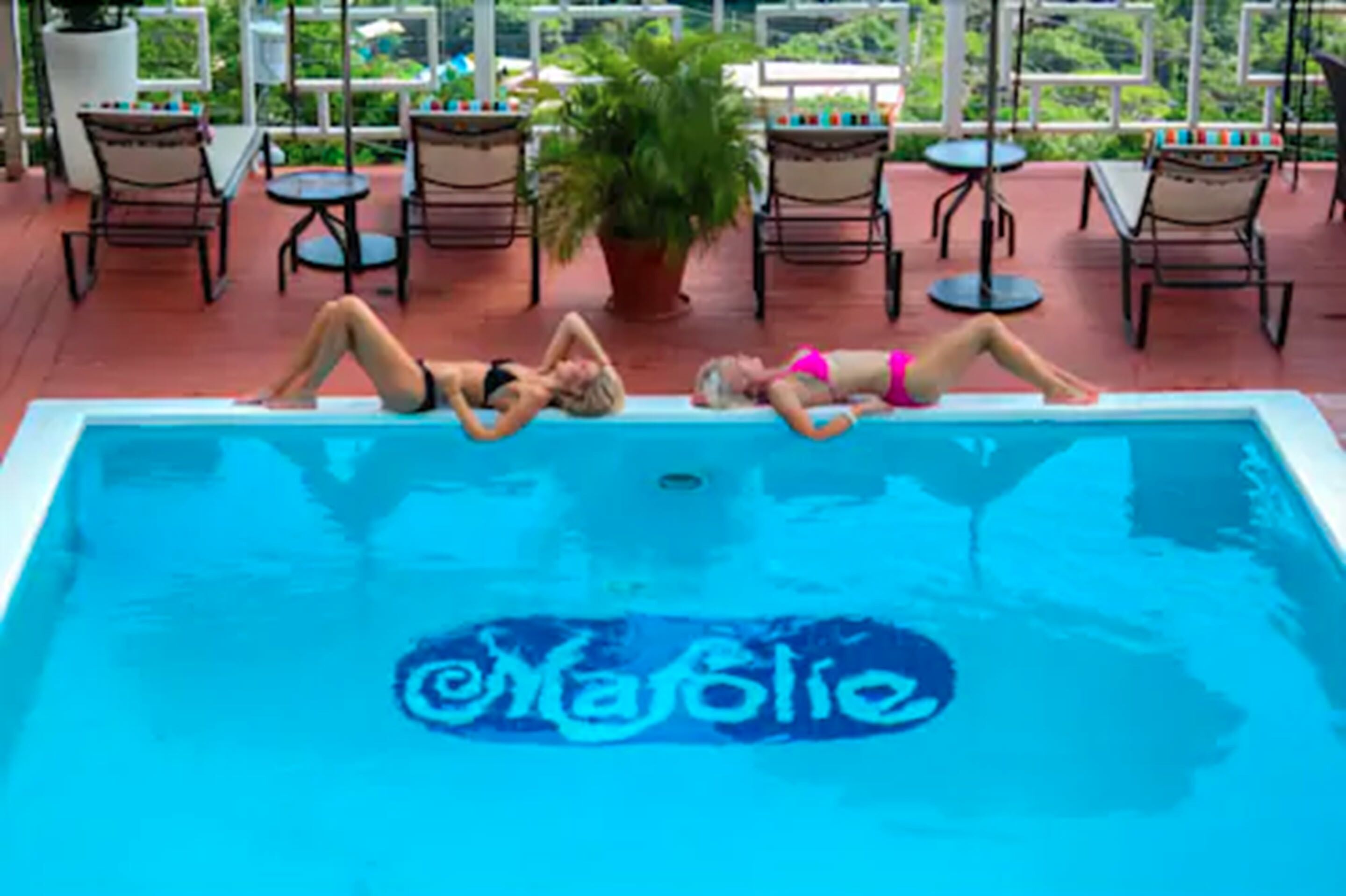 A swimming pool with three people lying on it, surrounded by lounge chairs and a potted plant.