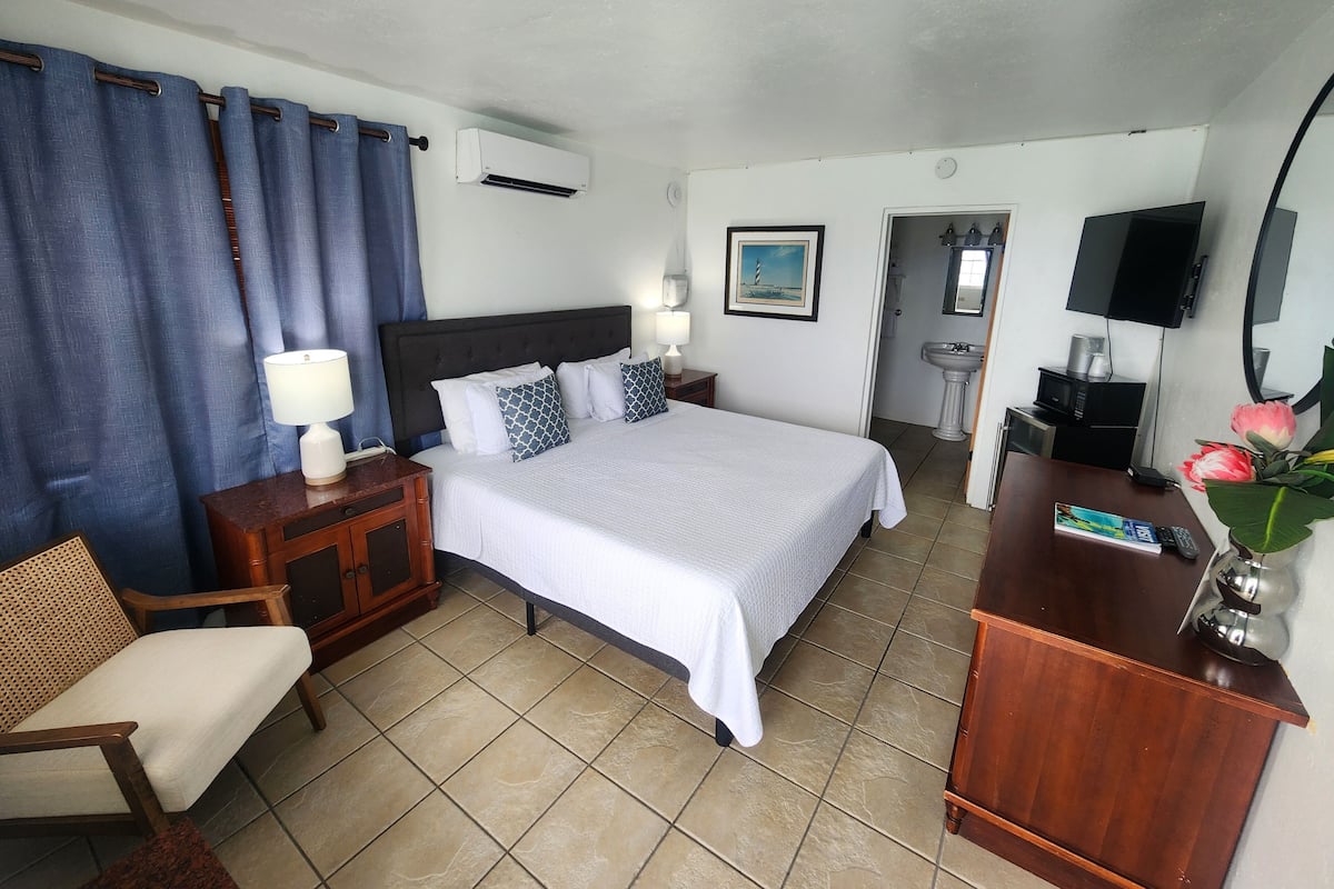 A hotel room with a bed, bedside tables, a chair, a desk, a television, and a bathroom visible through an open door.