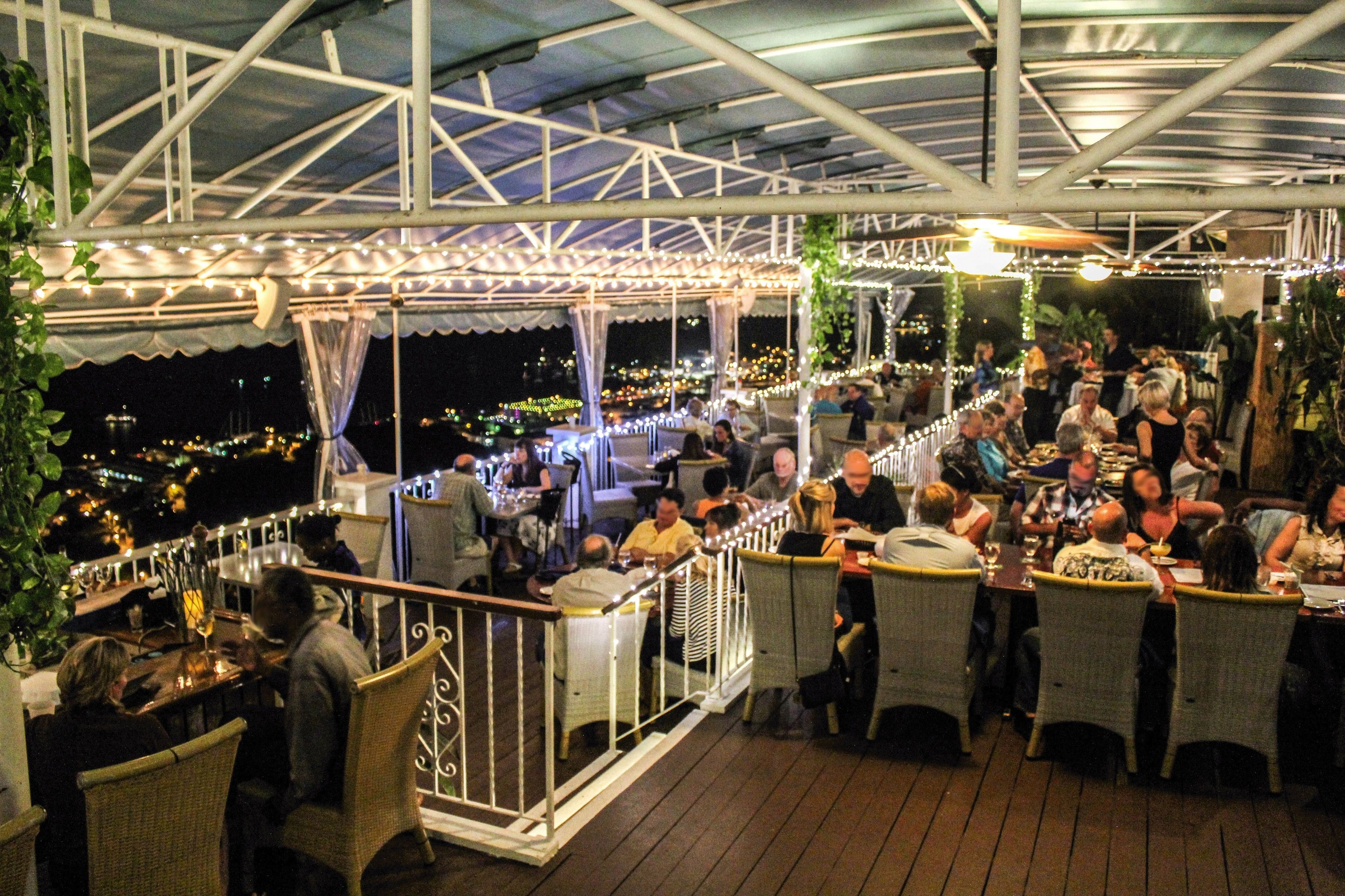 An outdoor dining area with tables and chairs, a covered roof, and a view of the city at night.