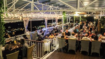 An outdoor dining area with tables and chairs, a covered roof, and a view of the city at night.