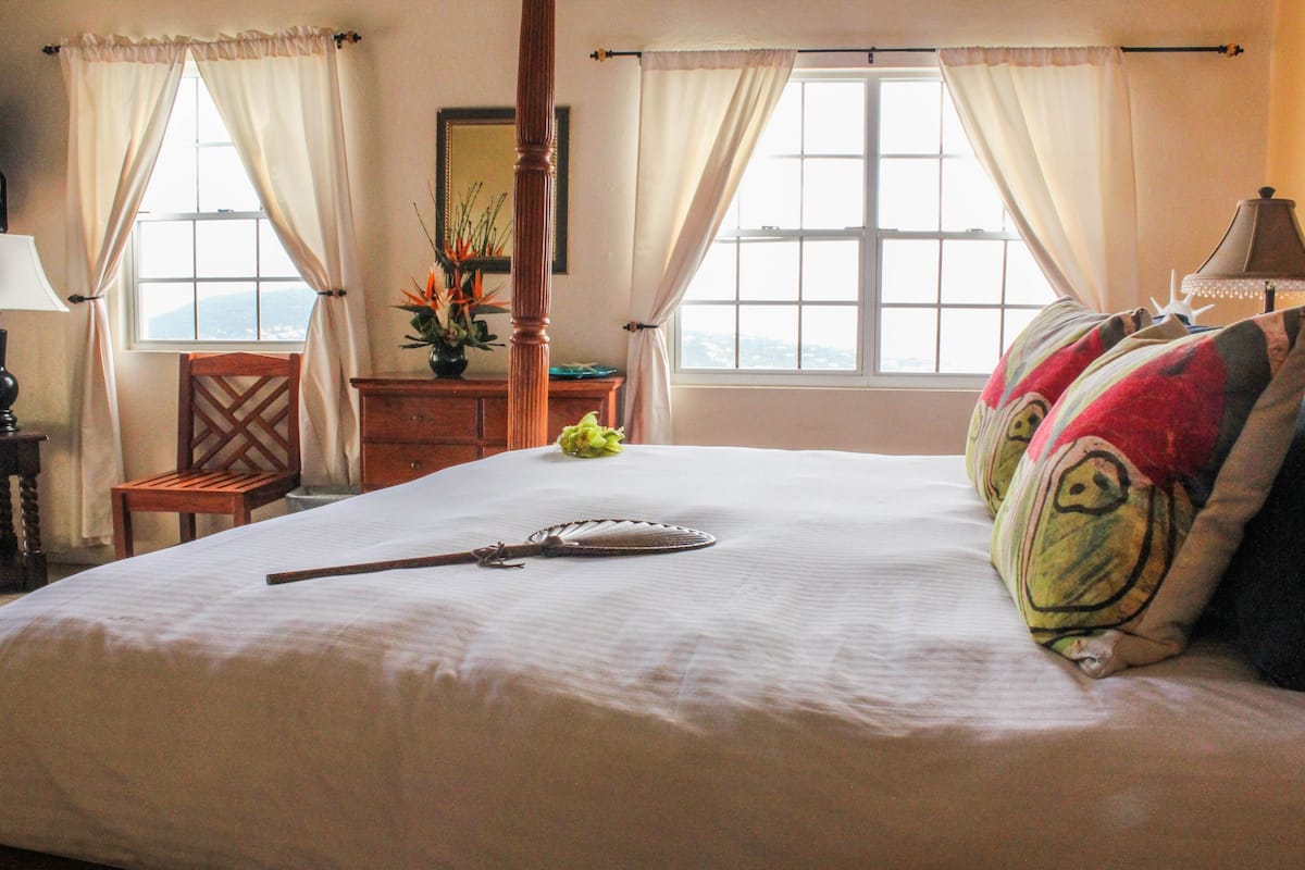 A neatly made bed with a decorative pillow, a wooden chair, a nightstand with a lamp, and a window with curtains.