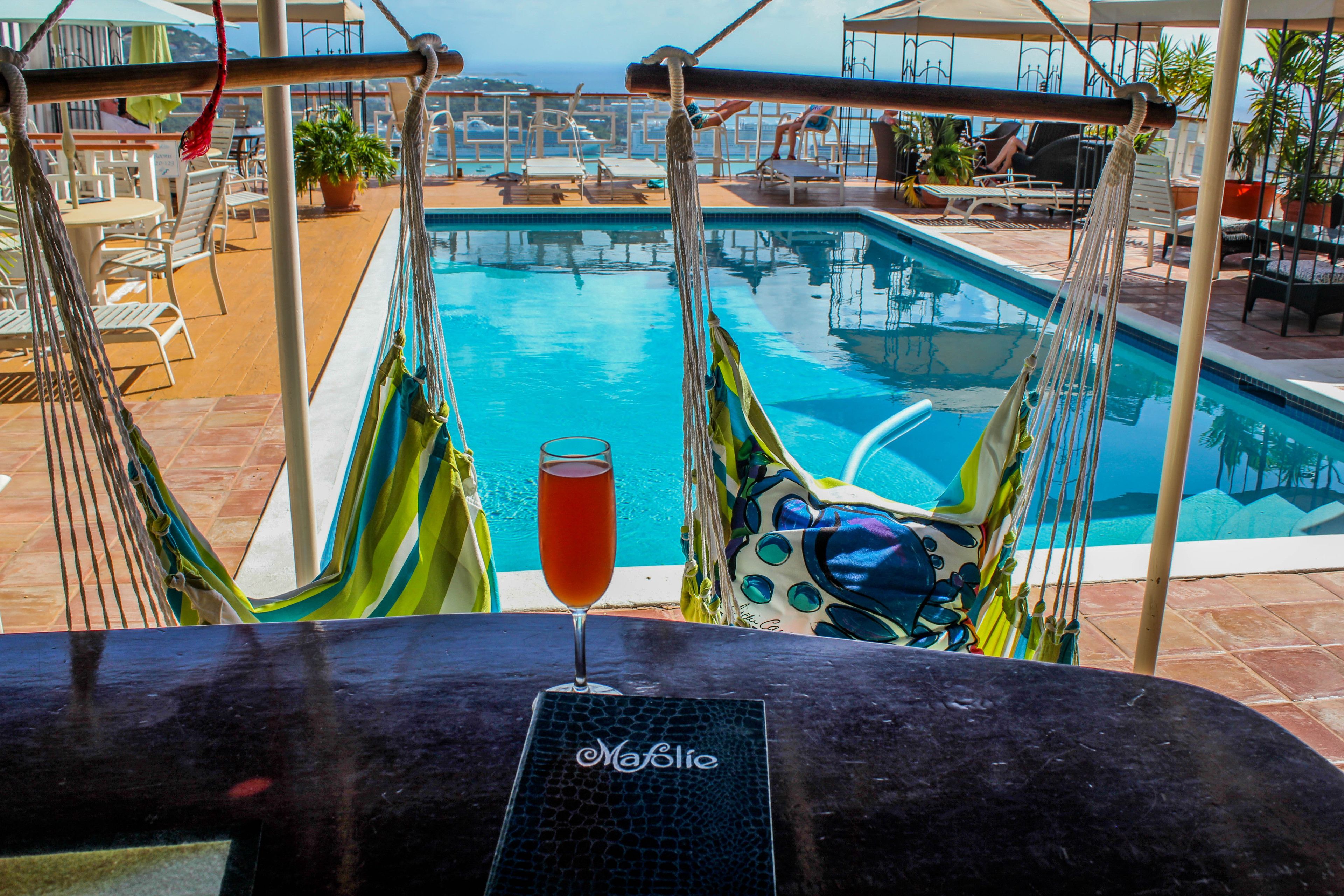 Vista de la piscina con una hamaca, sillones de descanso y una copa de vino tinto sobre una mesa.