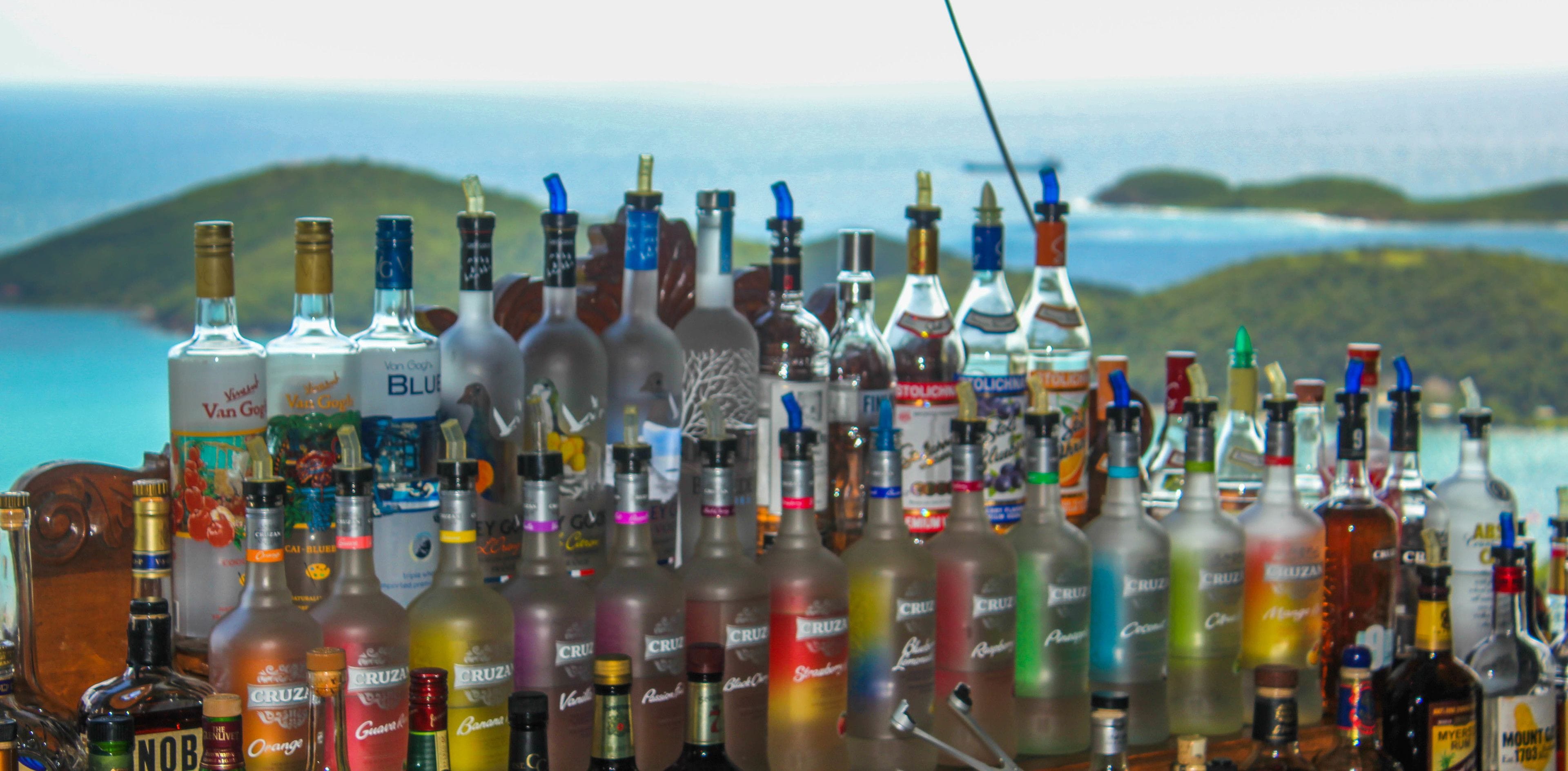 A collection of liquor bottles arranged on a counter with a scenic view of hills and water in the background.