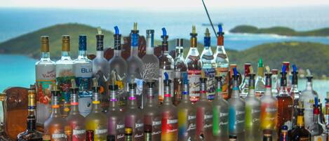 Varias botellas de licores dispuestas sobre una barra, con una vista panorámica de colinas y agua al fondo.