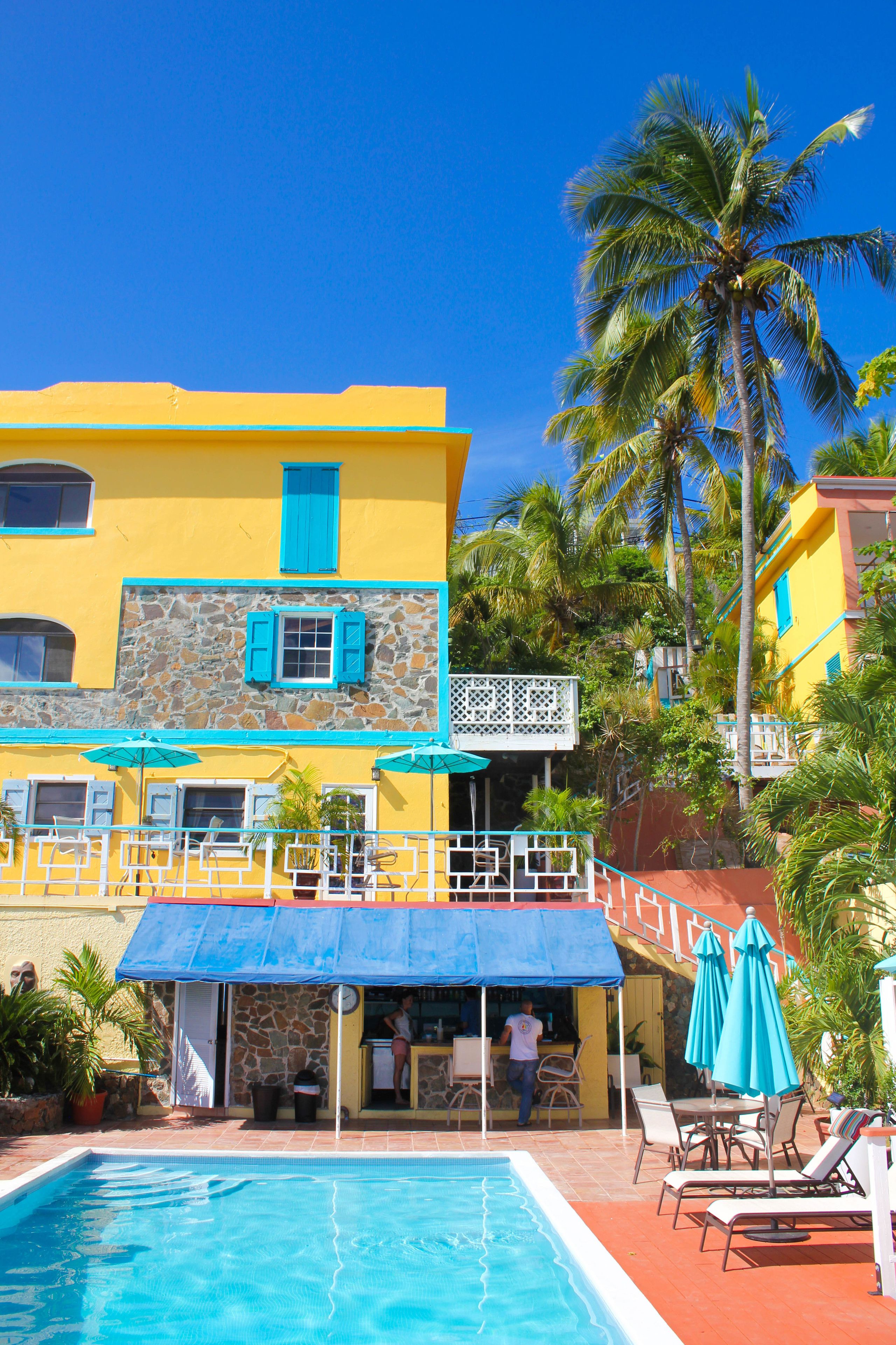 Un área junto a la piscina con un edificio amarillo, un muro de piedra y una palmera.