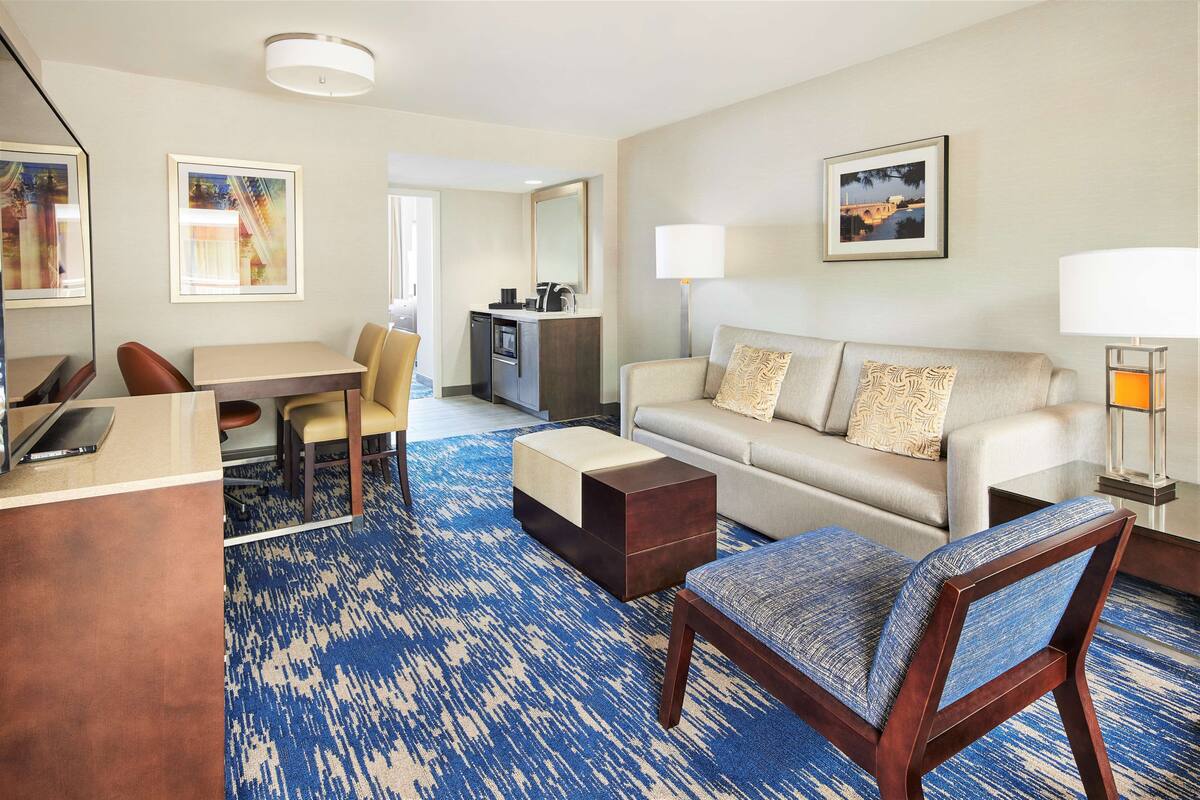 A hotel room with a sofa, armchair, dining table, and a small kitchenette.