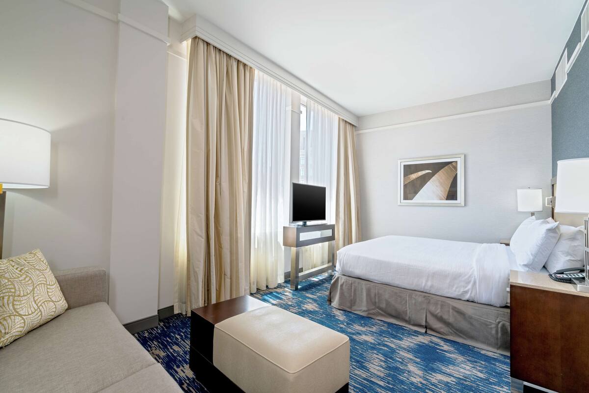 A hotel room with a bed, a sofa, a TV, a desk, and a window with curtains.