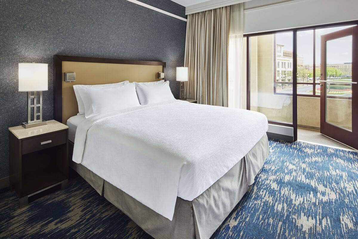 A hotel room with a large bed, two bedside tables with lamps, a patterned carpet, and a view through a large window.