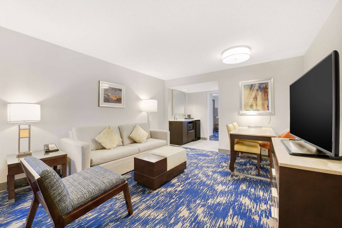 A hotel room with a sofa, armchair, coffee table, TV, desk, and lamp.