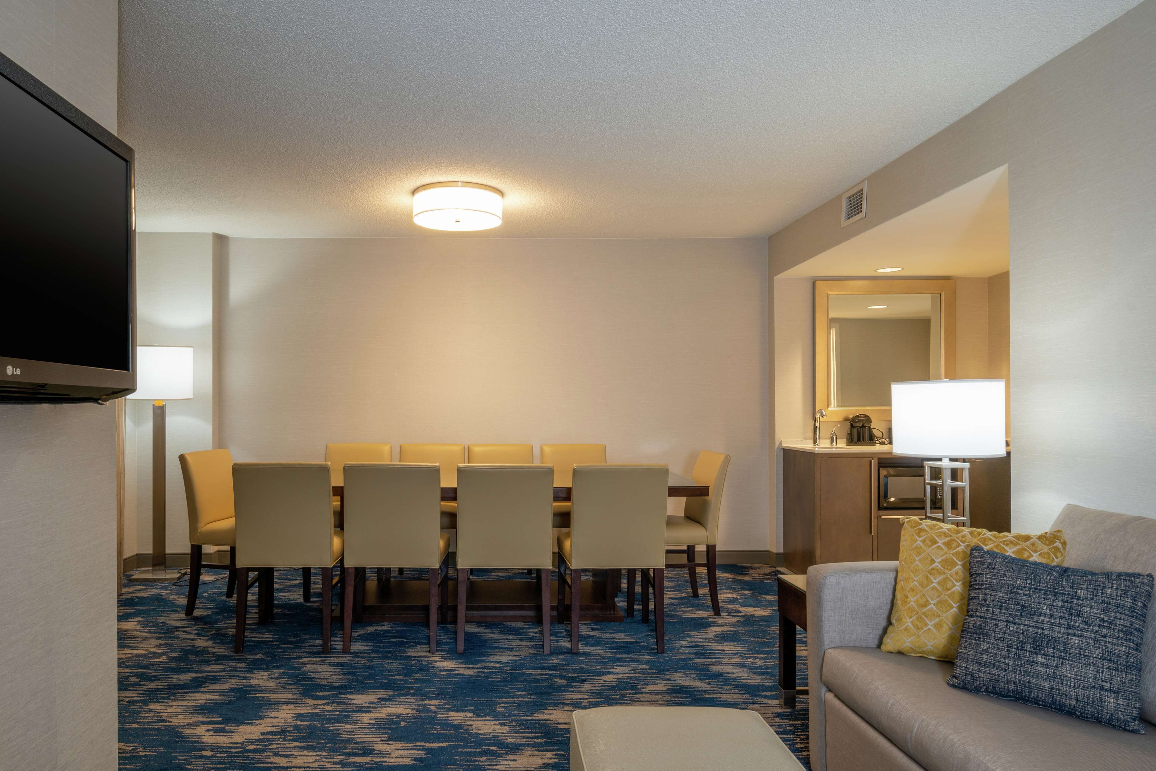 Habitación de hotel con zona de comedor, televisión de pantalla plana, sofá con cojines y alfombra azul estampada.