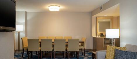 Quarto de hotel com área de jantar, televisão de tela plana, sofá com almofadas e carpete azul estampado.
