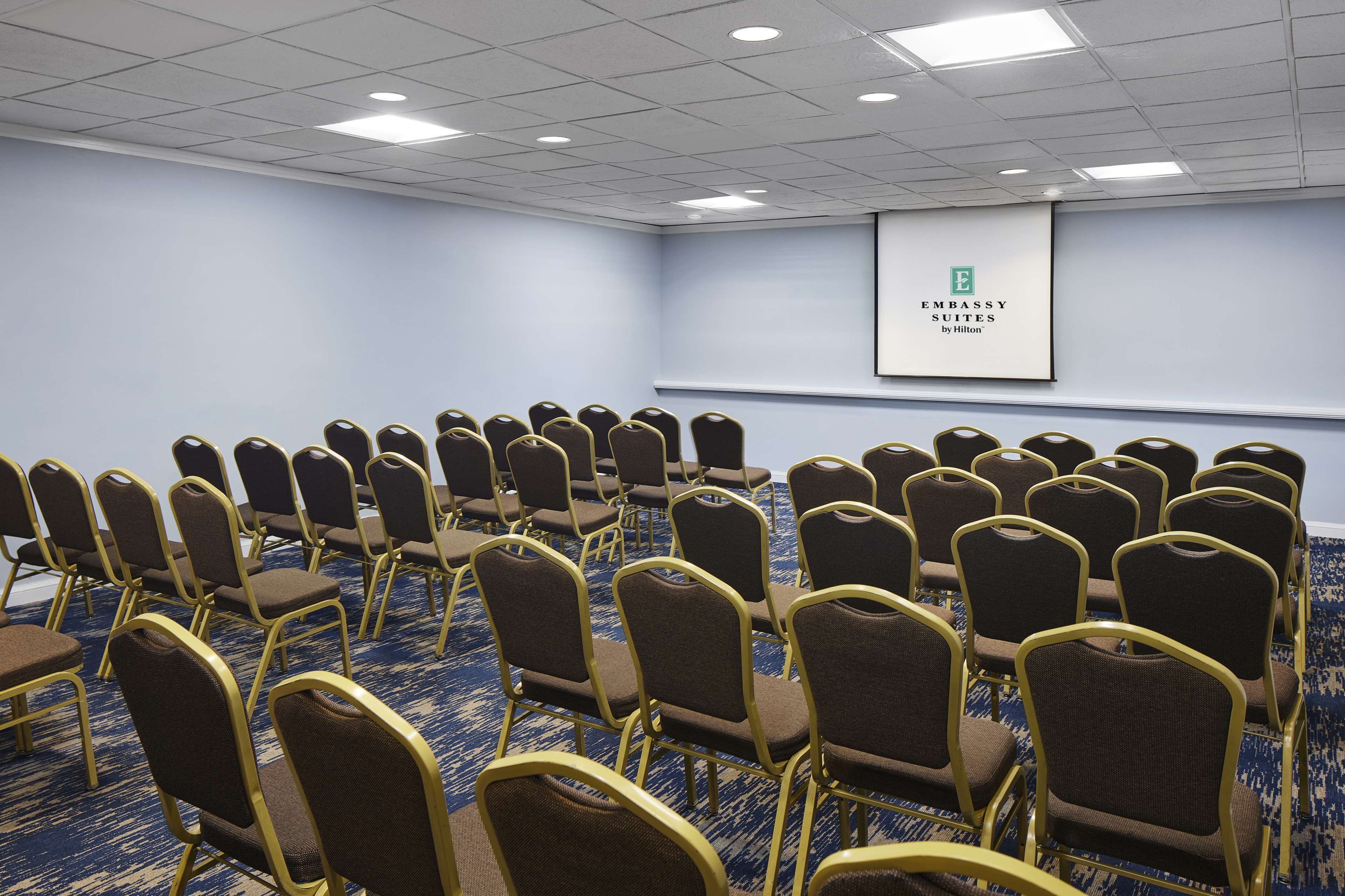 Un salón de conferencias con filas de sillas, una pantalla de proyector mostrando 'Embassy Suites by Hilton' y piso alfombrado.