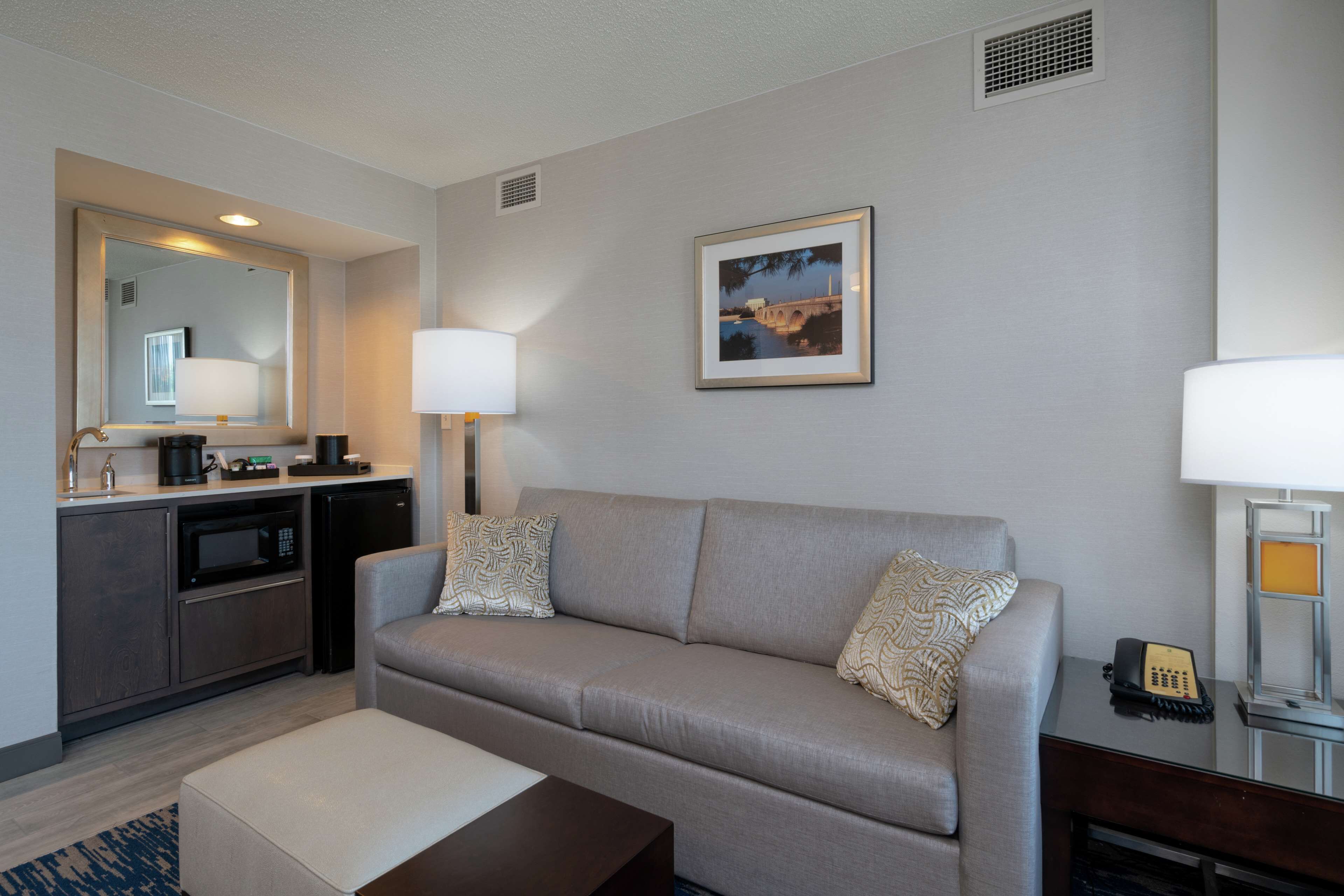 A modern hotel room with a sofa, a coffee table, a desk with a telephone, a lamp, a microwave, and a small kitchen area.