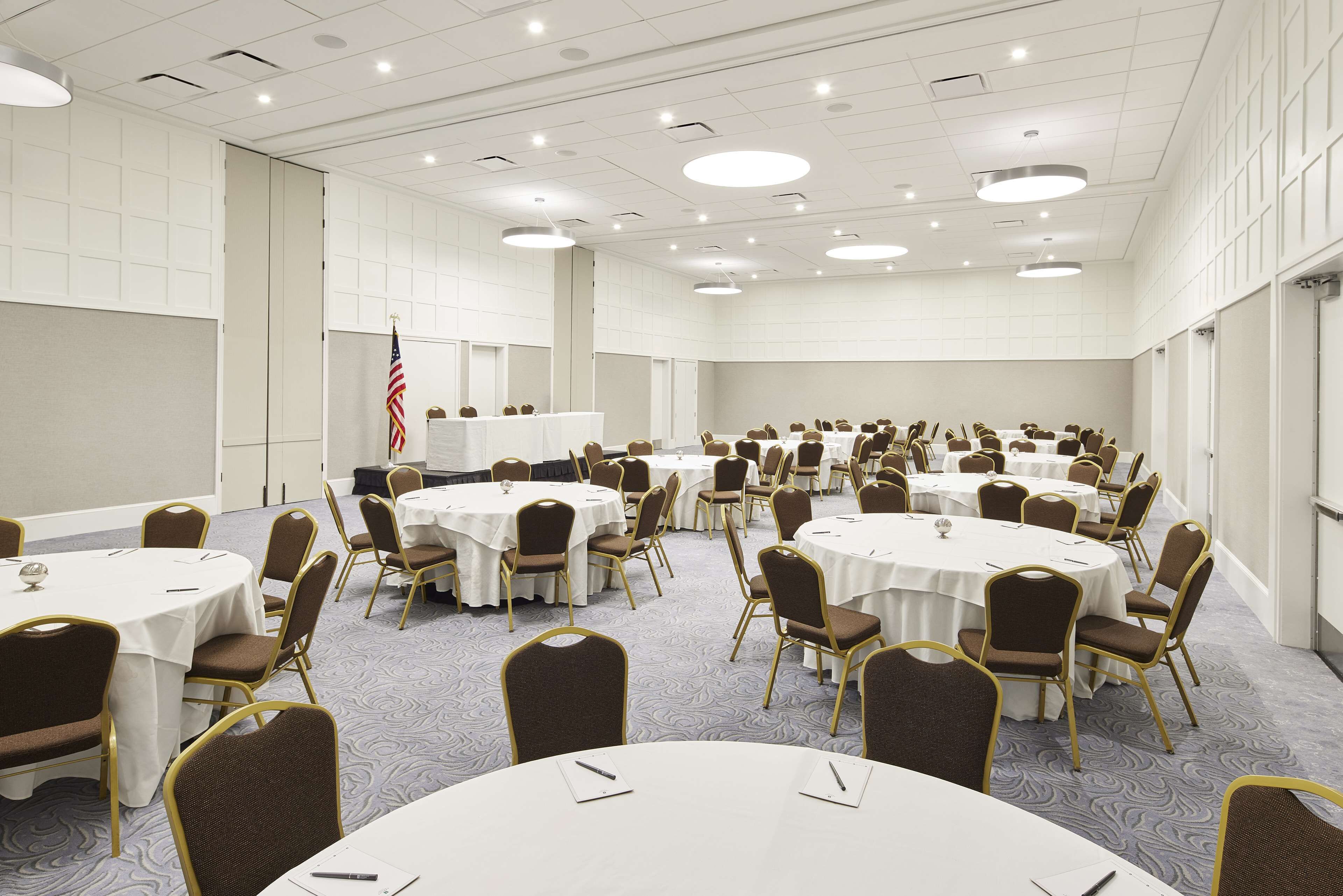 Un salón de conferencias con mesas y sillas redondas, paredes blancas y suelo alfombrado.