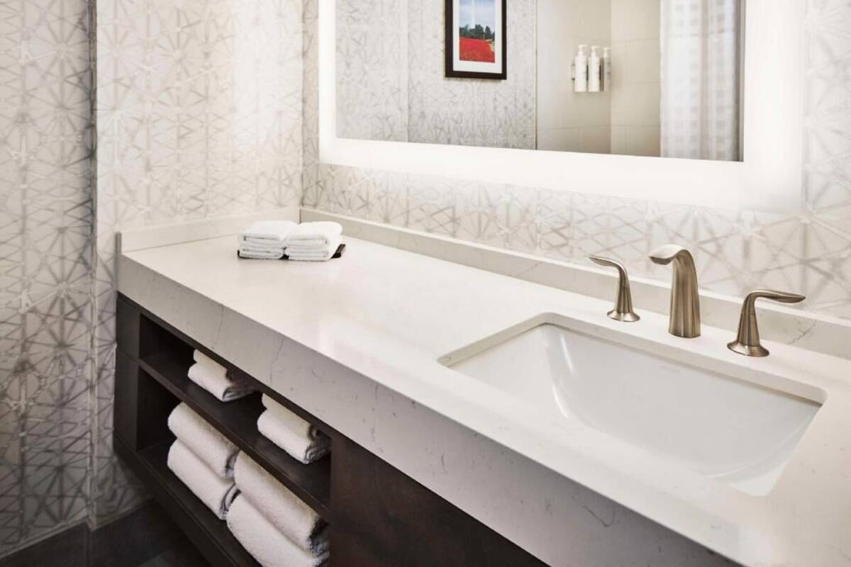 A modern bathroom with a large sink, two faucets, and a mirror above it. There are towels on a shelf below the sink.