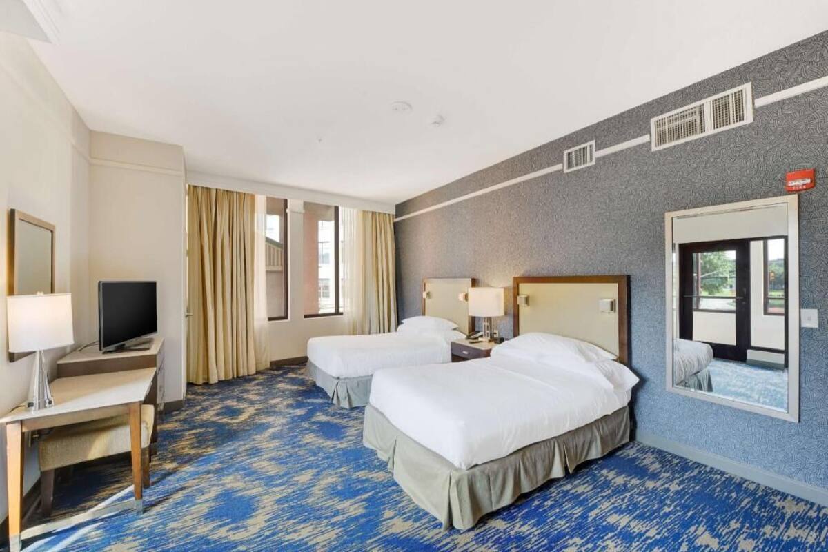 A hotel room with two beds, a desk with a TV, a window with curtains, and a patterned carpet.