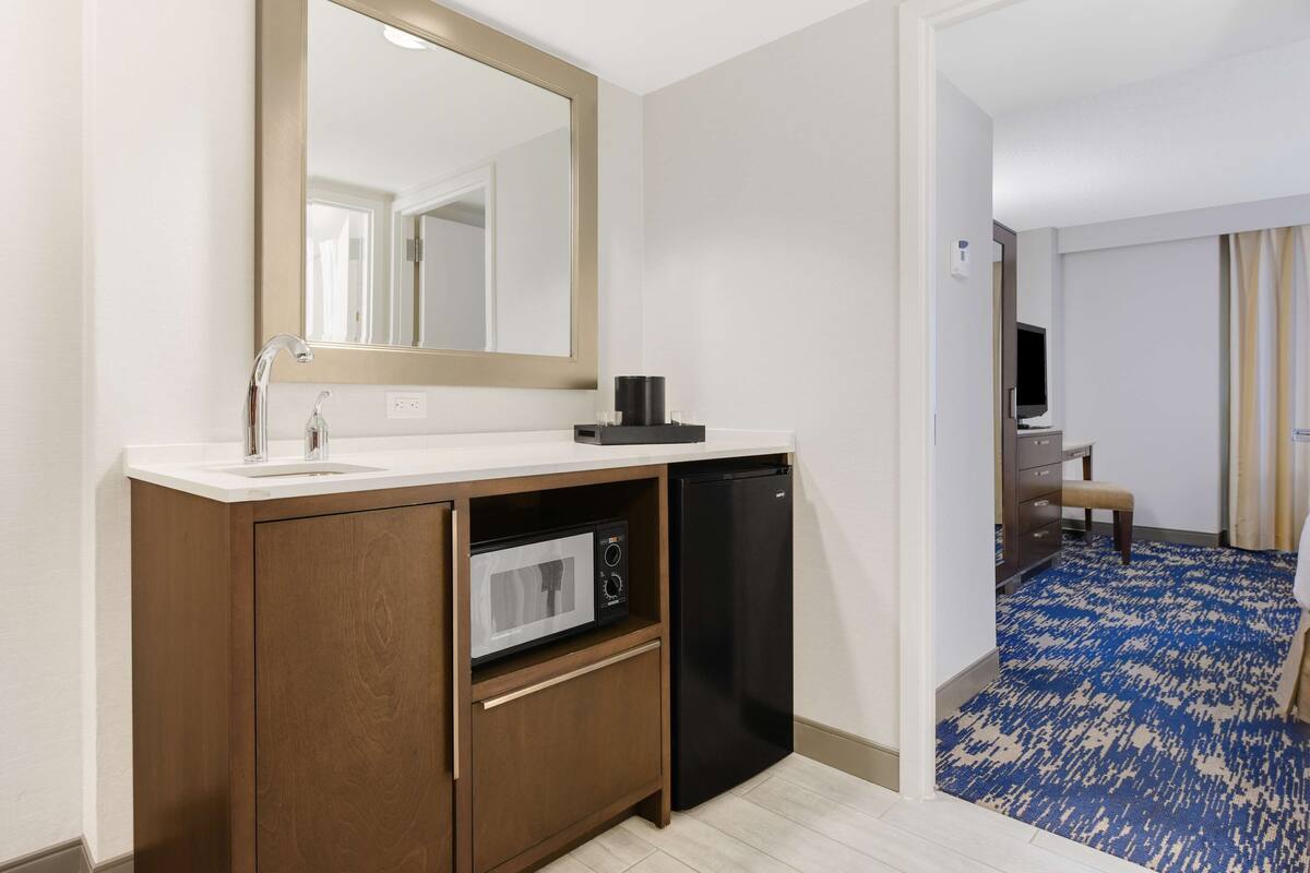 A hotel room with a wooden cabinet, a microwave, a black refrigerator, a white sink, and a large mirror.