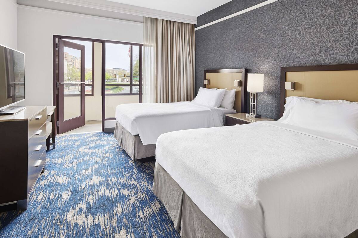 A hotel room with two beds, a TV, a desk, and a view of the city.