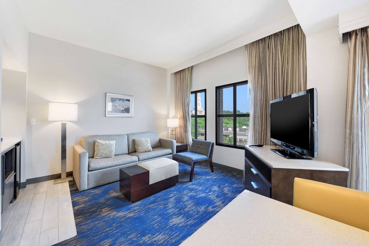 A hotel room with a sofa, armchair, TV, and a view of a building through the window.