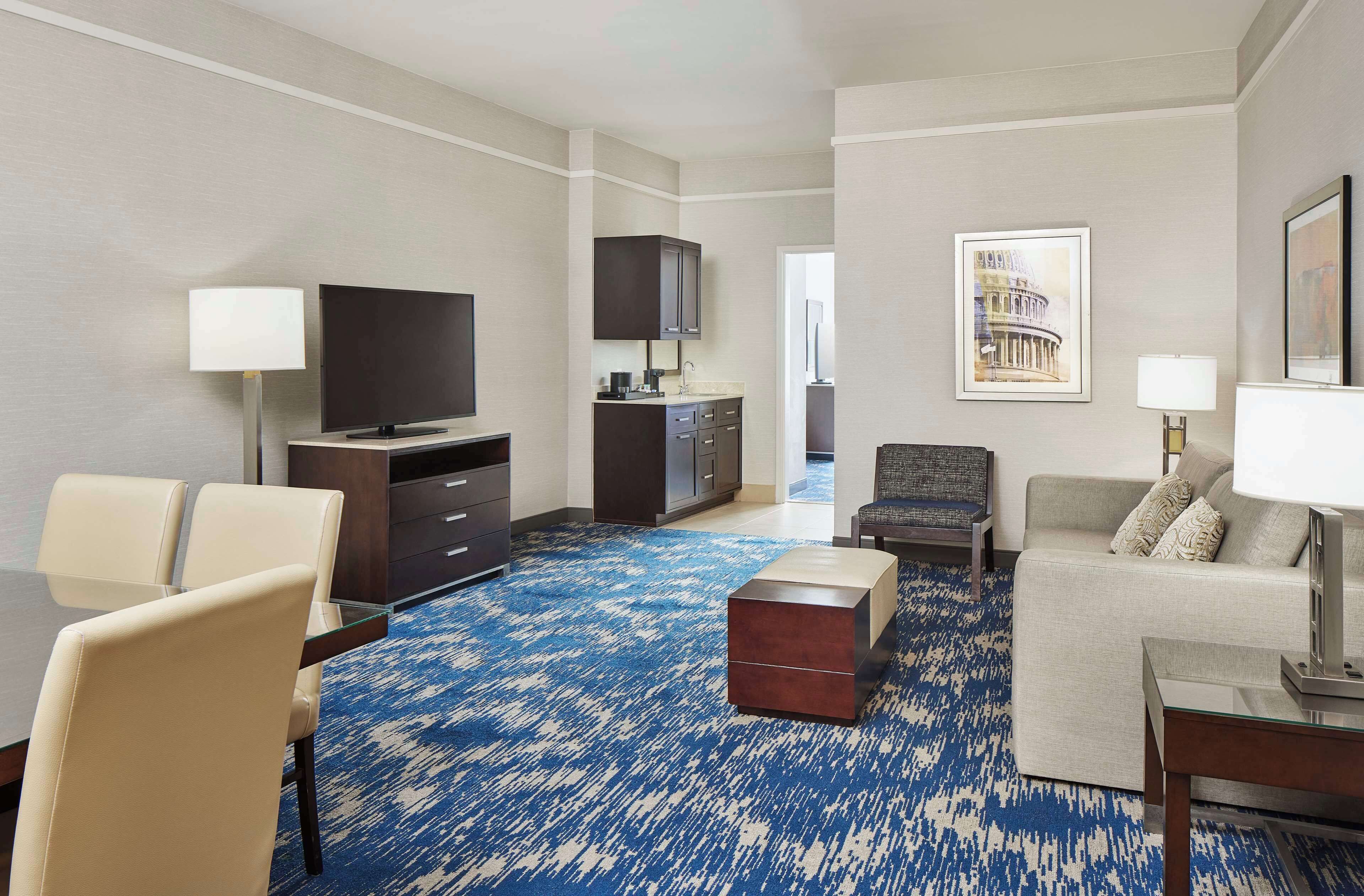 A hotel room with a flat-screen TV, a sofa, a dining table with chairs, a coffee machine, and a framed picture on the wall.