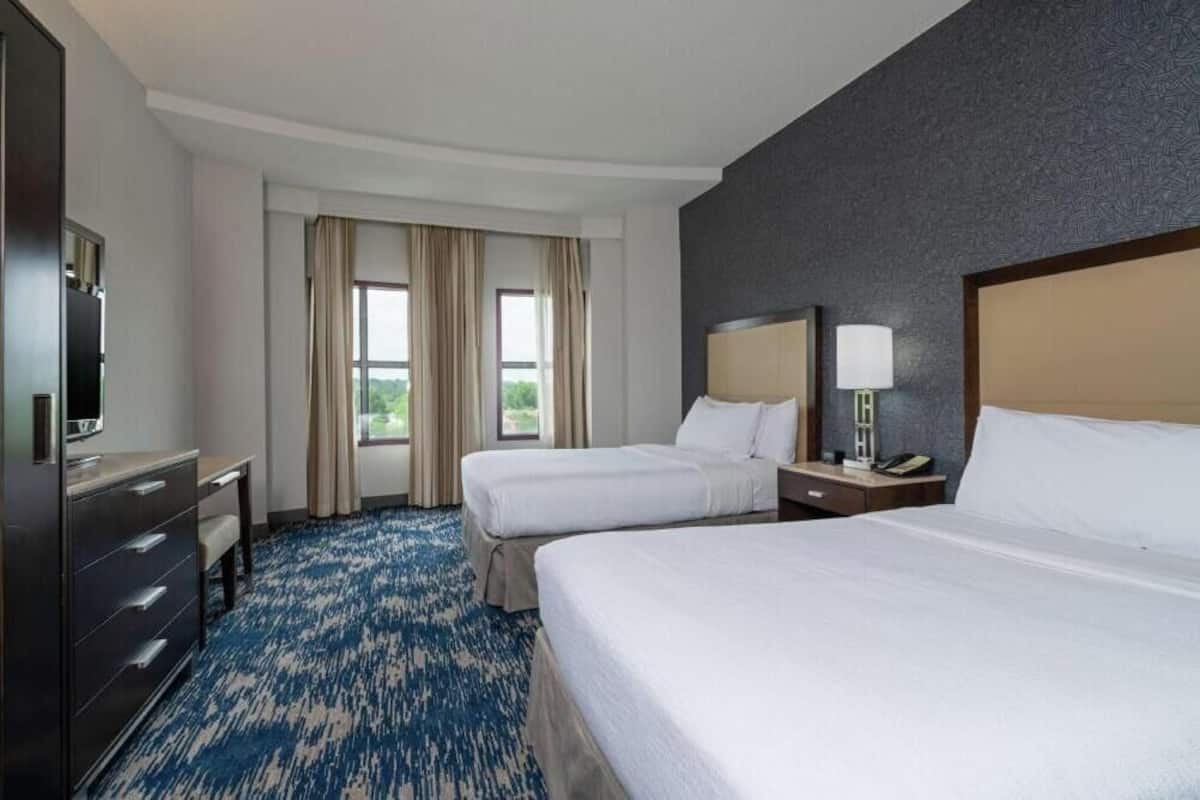 A hotel room with two beds, a desk, a dresser, a TV, and a window with curtains.