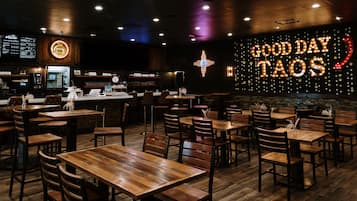Interior de un restaurante con mesas y sillas de madera, una barra con máquina de café, y una pared con el letrero iluminado 'GOOD DAY TAOS'.