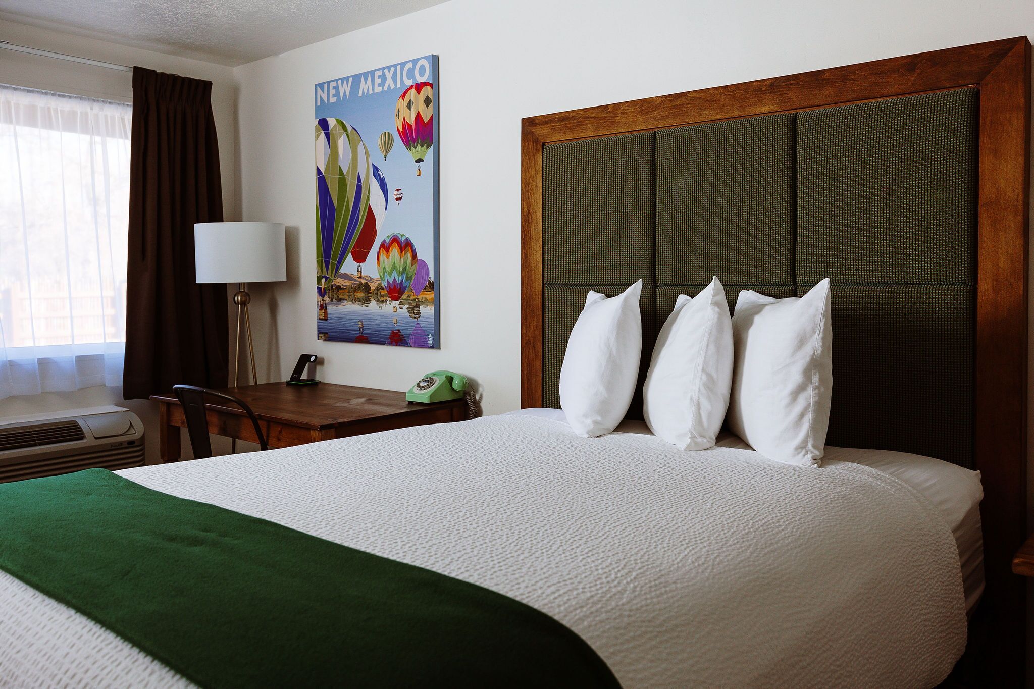 A hotel room with a large bed, a wooden headboard, a green throw blanket, a wall art of hot air balloons, a desk with a lamp, and a telephone.