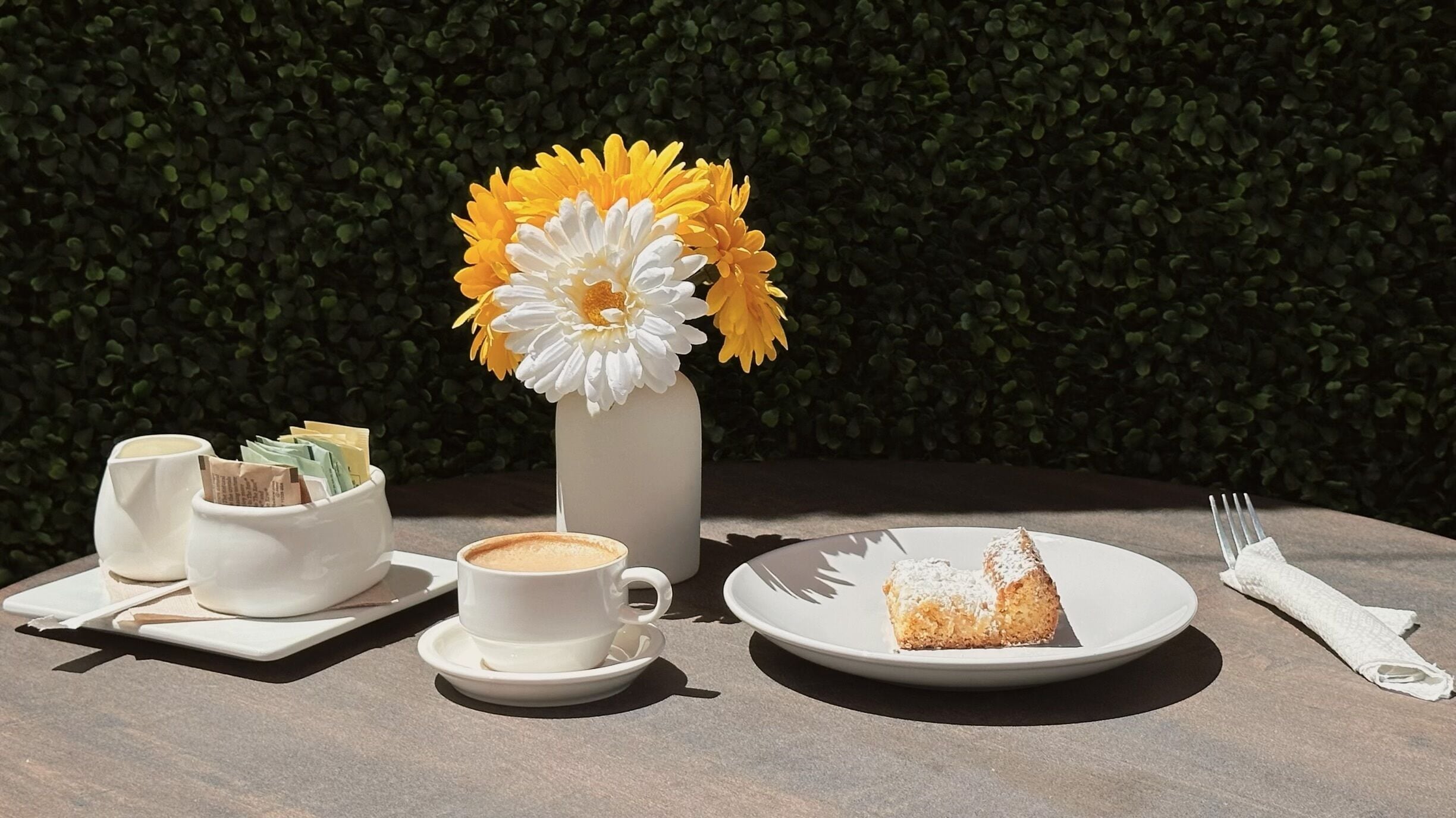 A table with a cup of coffee, a plate with a pastry, a vase with flowers, and a napkin.