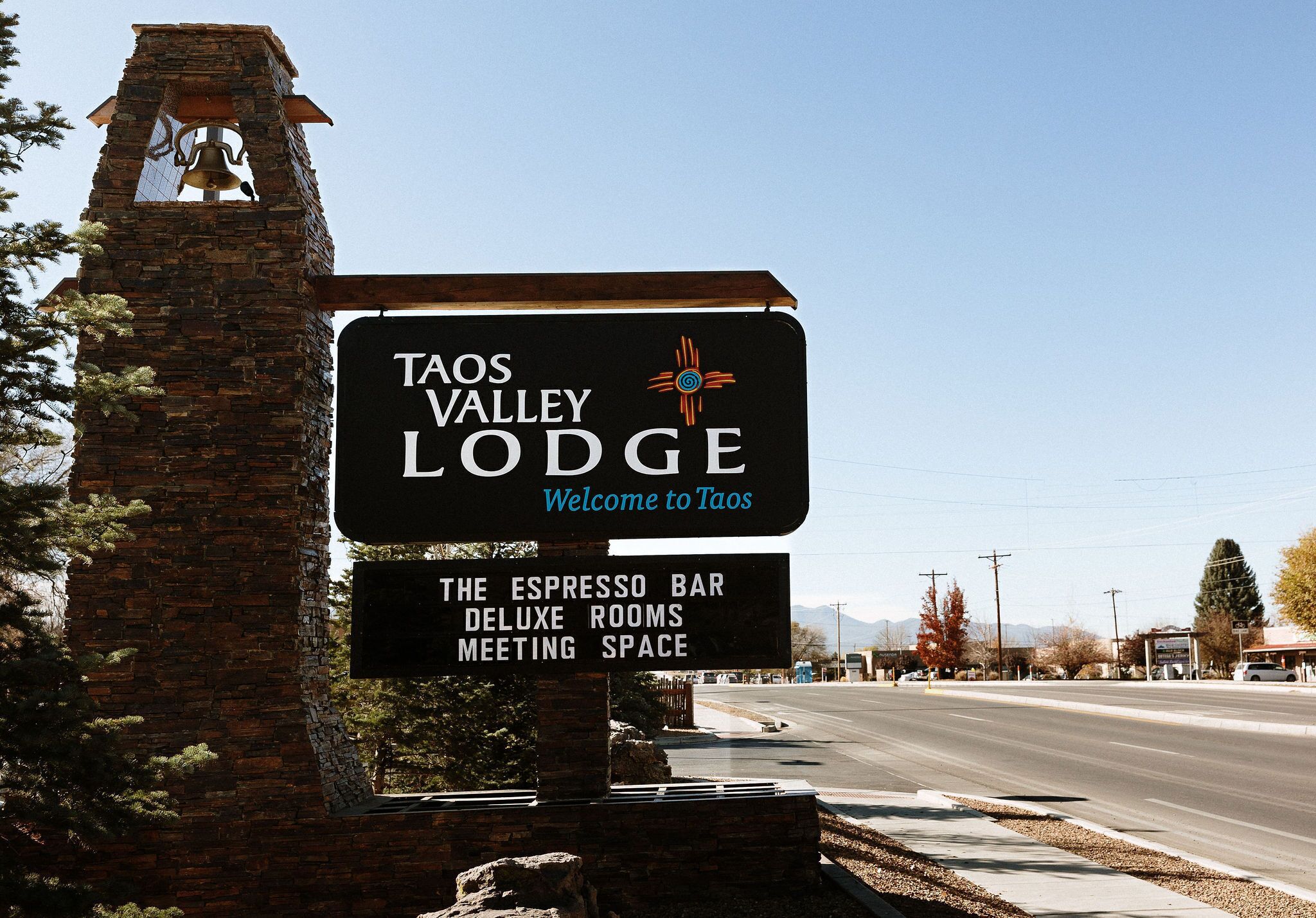 Señal de Taos Valley Lodge con una torre de campanas, que indica una entrada acogedora y destaca servicios como cafetería con barra de expresos, habitaciones de lujo y salas para reuniones.