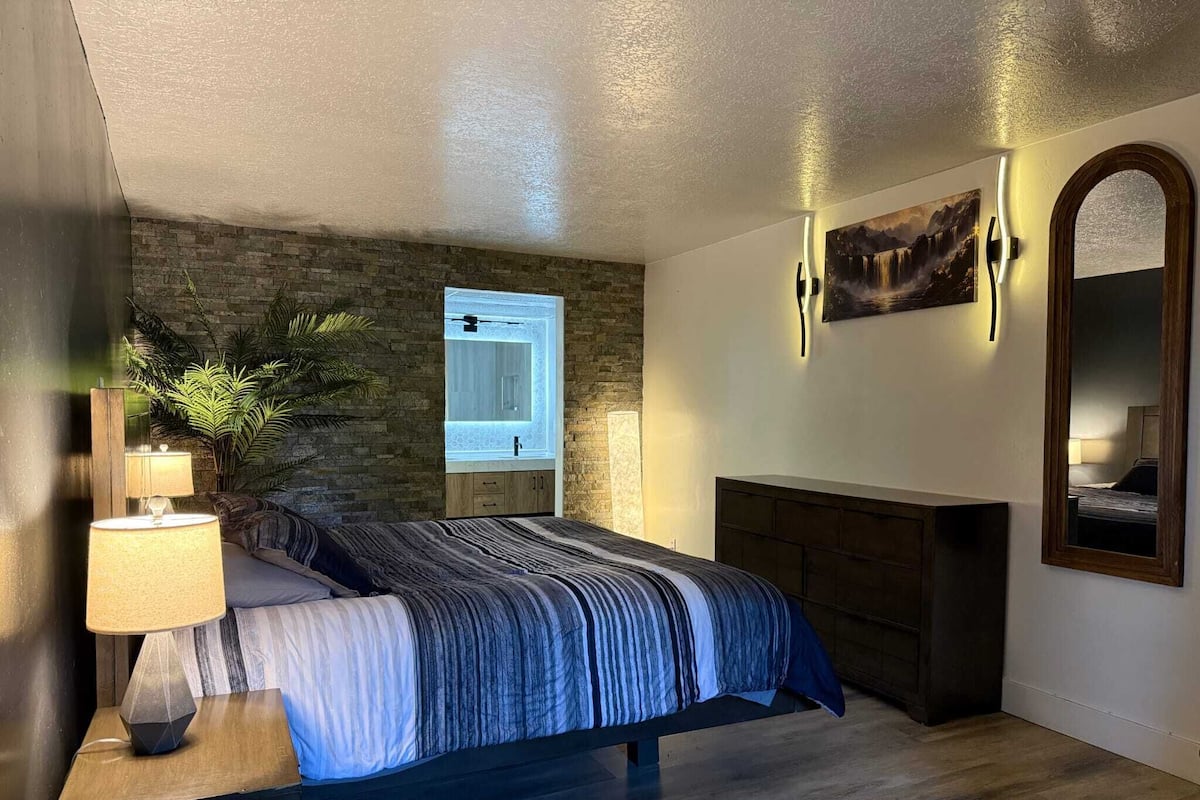 A bedroom with a stone wall, a bed with a striped blanket, a nightstand with a lamp, a window with a view of a mountain, and a fireplace.