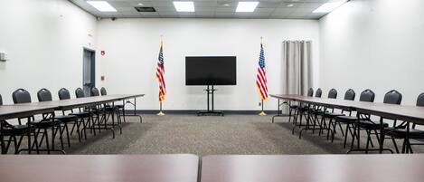 Un salón de conferencias con una larga mesa, filas de sillas, dos banderas estadounidenses, un televisor de pantalla plana y una puerta.