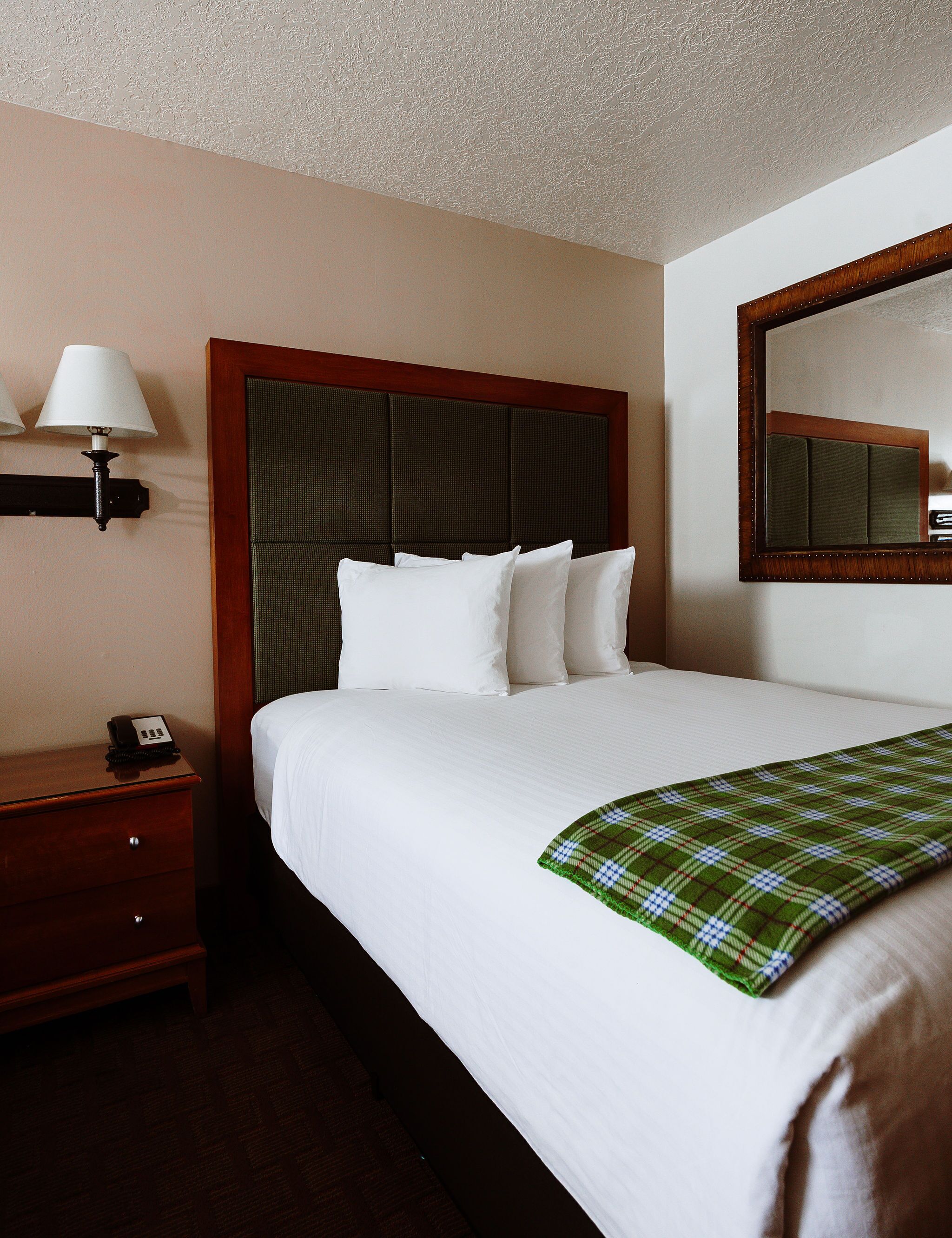 A hotel room with a neatly made bed, a bedside table with a lamp, a telephone, and a mirror on the wall.