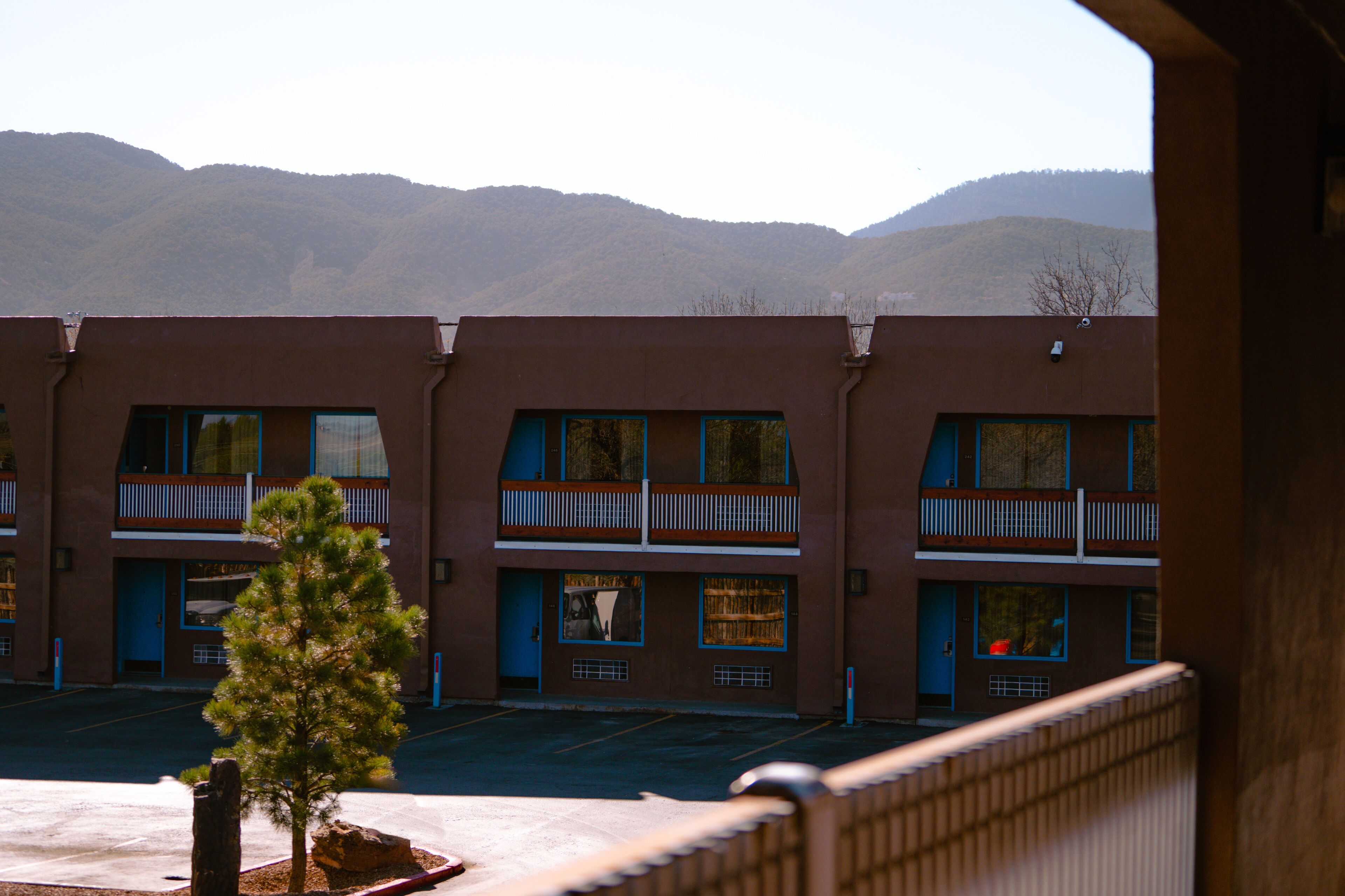 A row of two-story buildings with balconies, a parking lot, and mountains in the background.