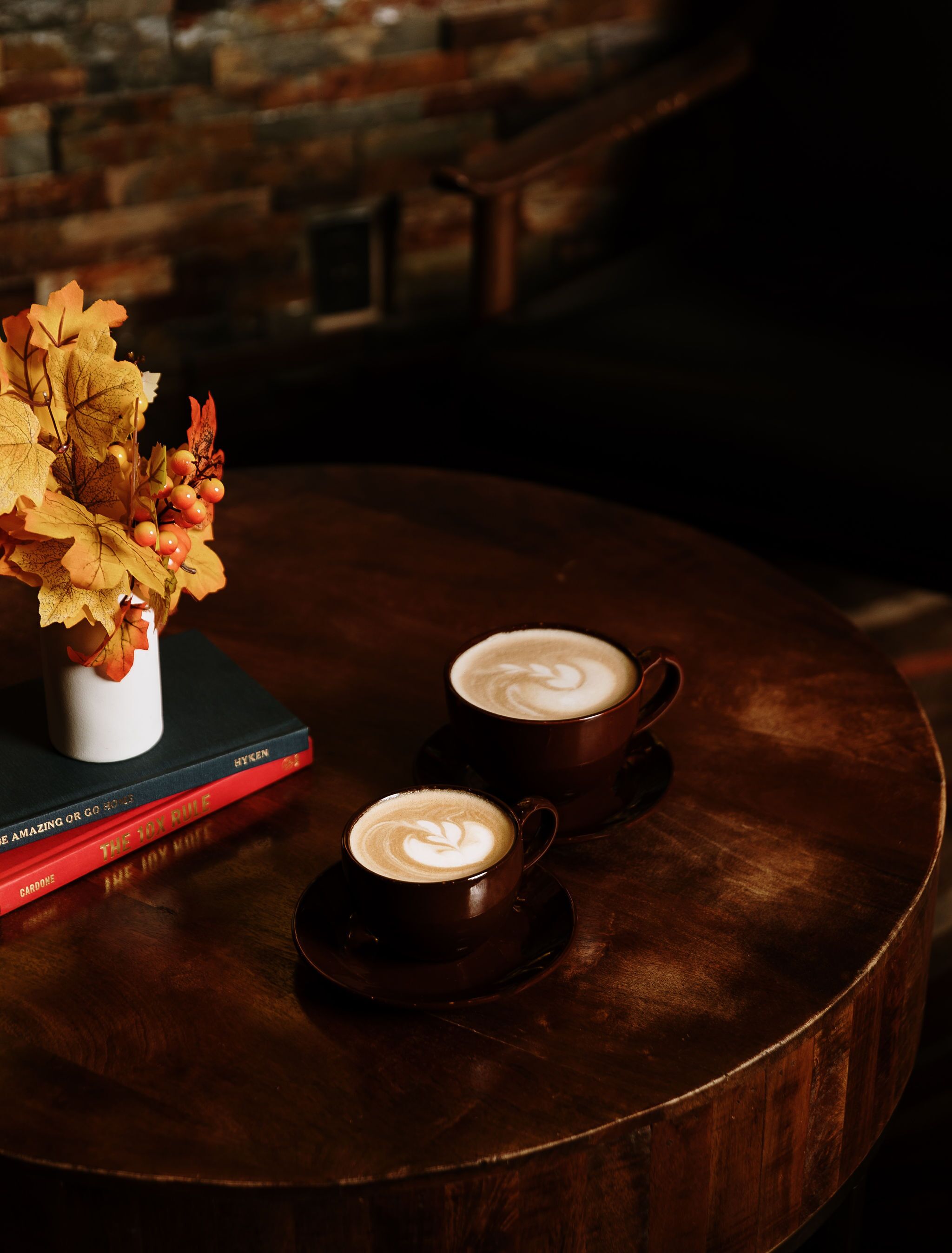 A round wooden table with two dark ceramic cups of coffee, a white vase with autumn leaves, and a stack of books.
