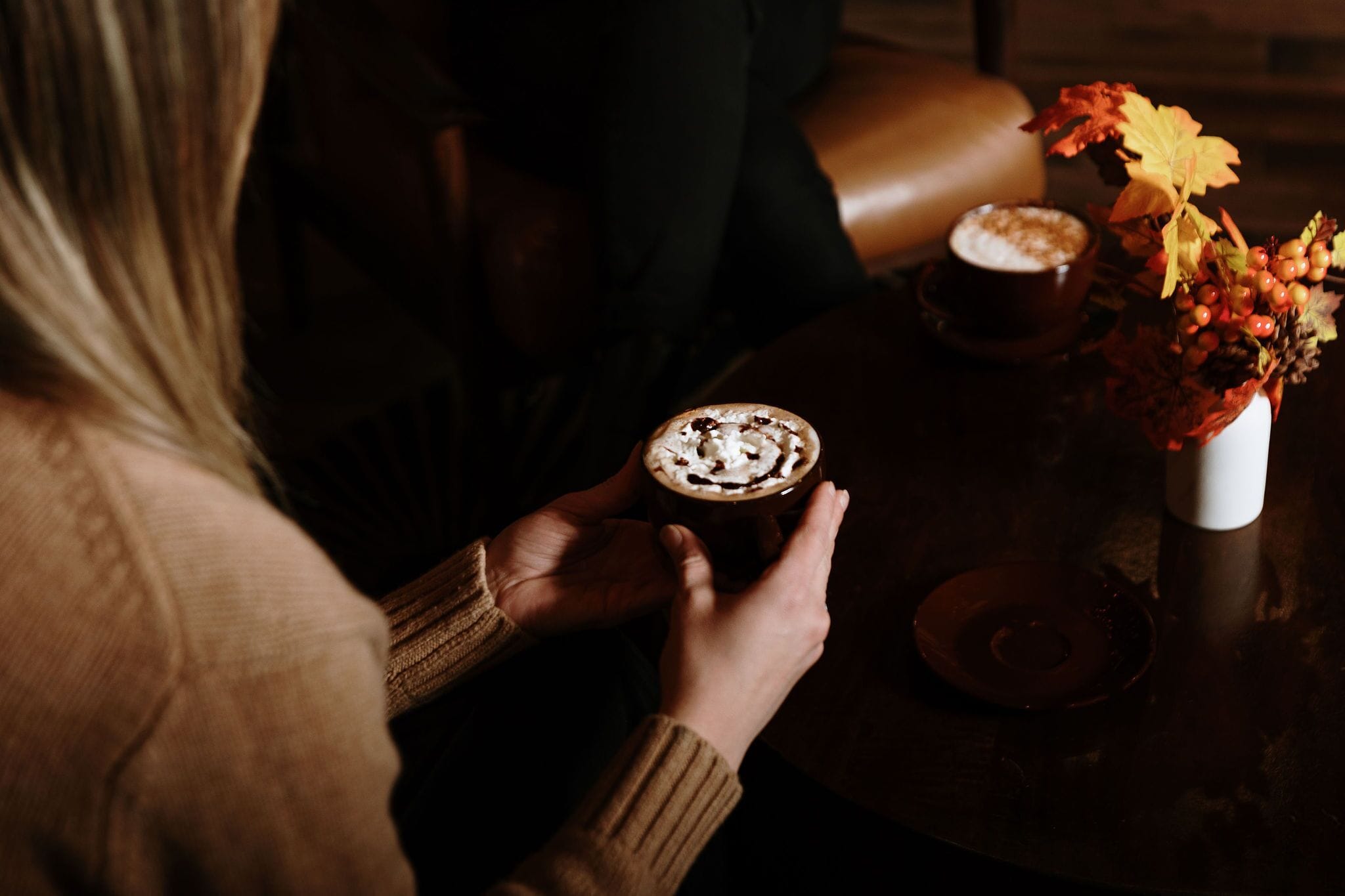 Una persona sosteniendo una taza de café con crema batida y virutas de chocolate, sentada a una mesa con un jarrón de hojas otoñales.