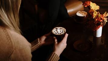 Una persona sosteniendo una taza de café con crema batida y virutas de chocolate, sentada a una mesa con un jarrón de hojas otoñales.