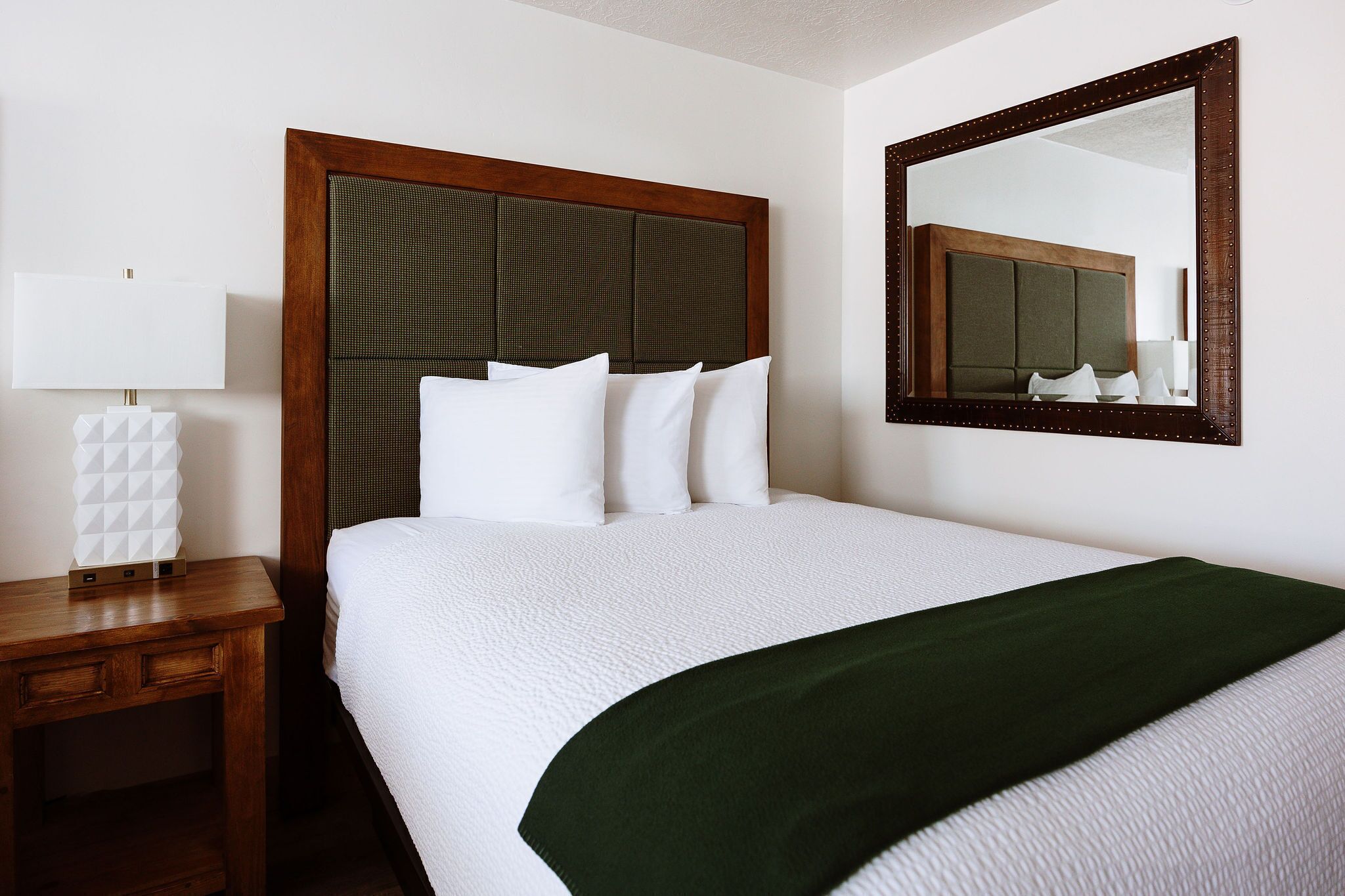 A neatly made bed with white bedding and a green throw, a wooden nightstand with a lamp, and a large mirror on the wall.