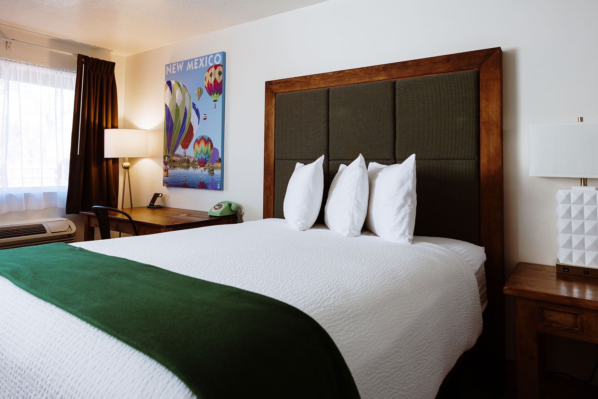 A hotel room with a large bed, a green throw blanket, a wall-mounted lamp, a telephone, and a poster of New Mexico.