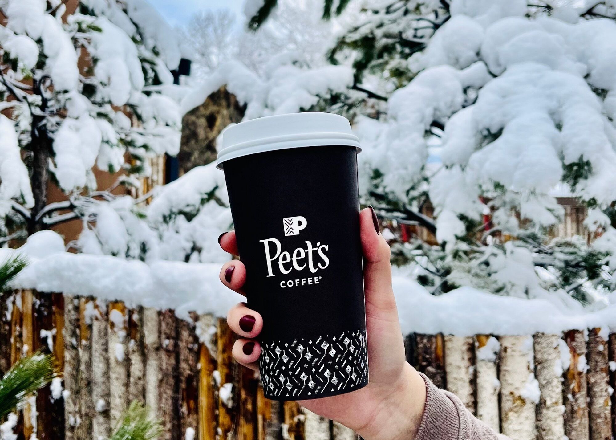 A hand holding a black coffee cup with white text and a logo, against a snowy background with a wooden fence.