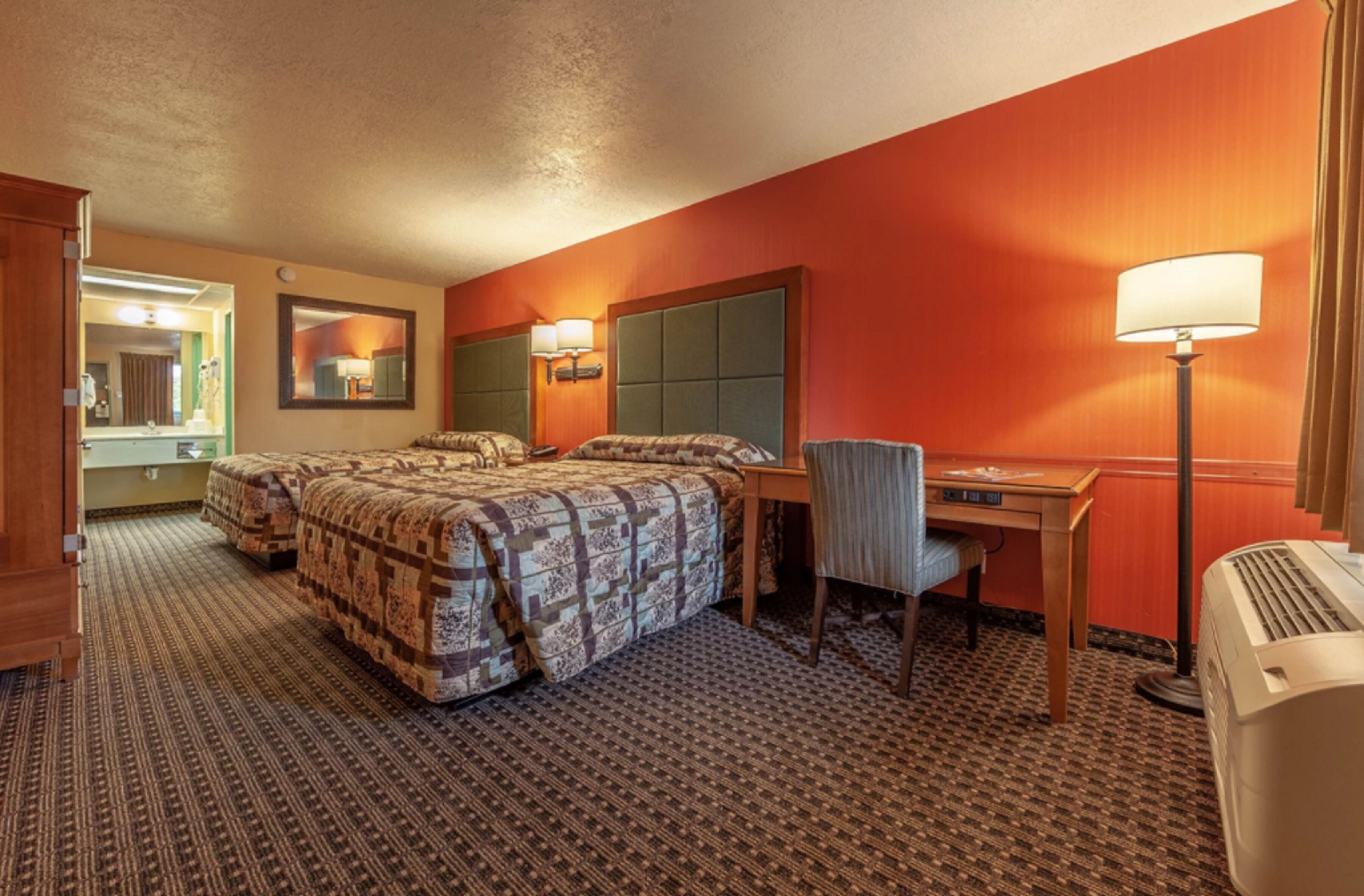 A hotel room with two beds, a desk, a chair, a lamp, and a heater.
