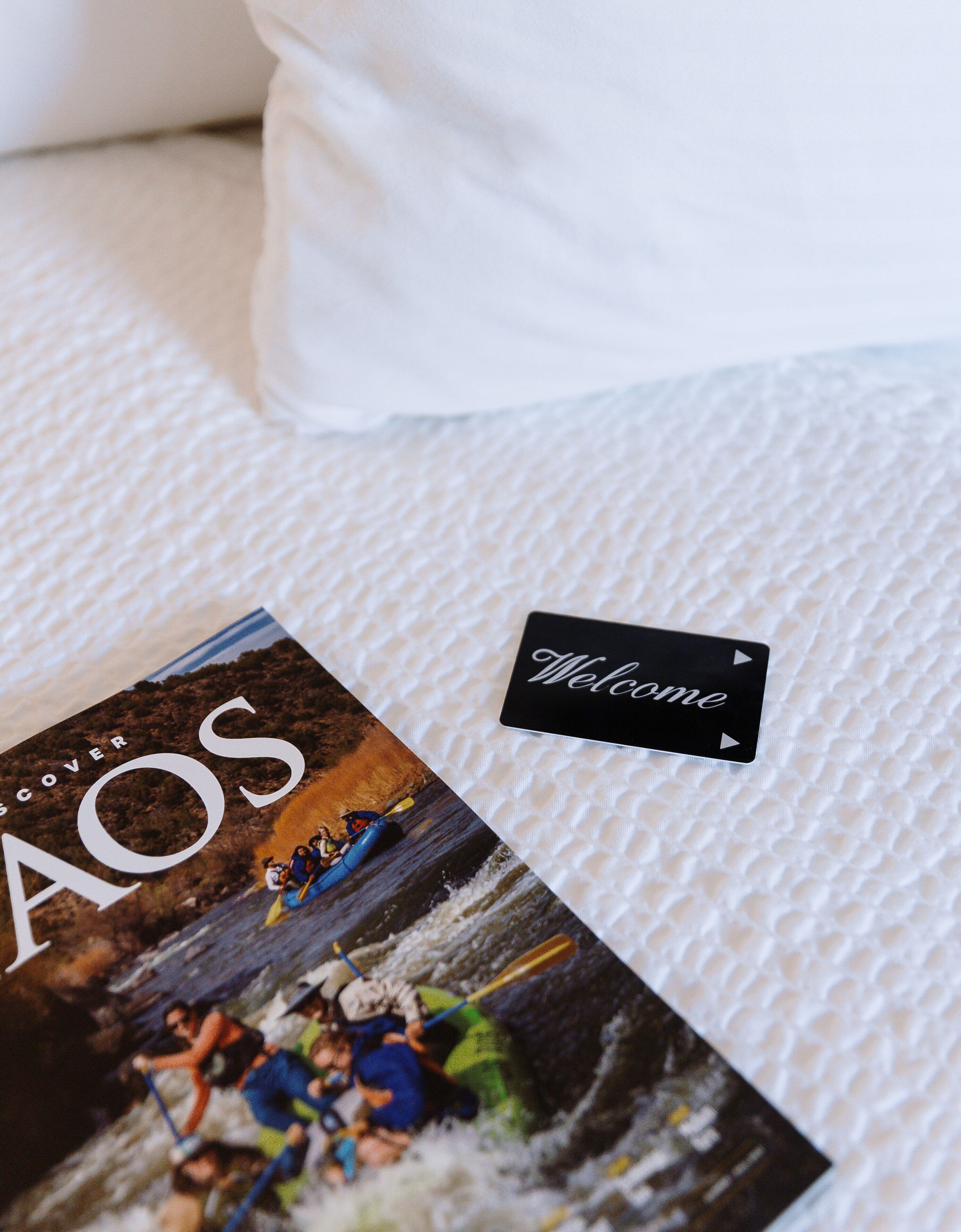 A bed with a white textured cover, a welcome card, and a magazine titled 'Discover AOS' featuring a kayaking scene.