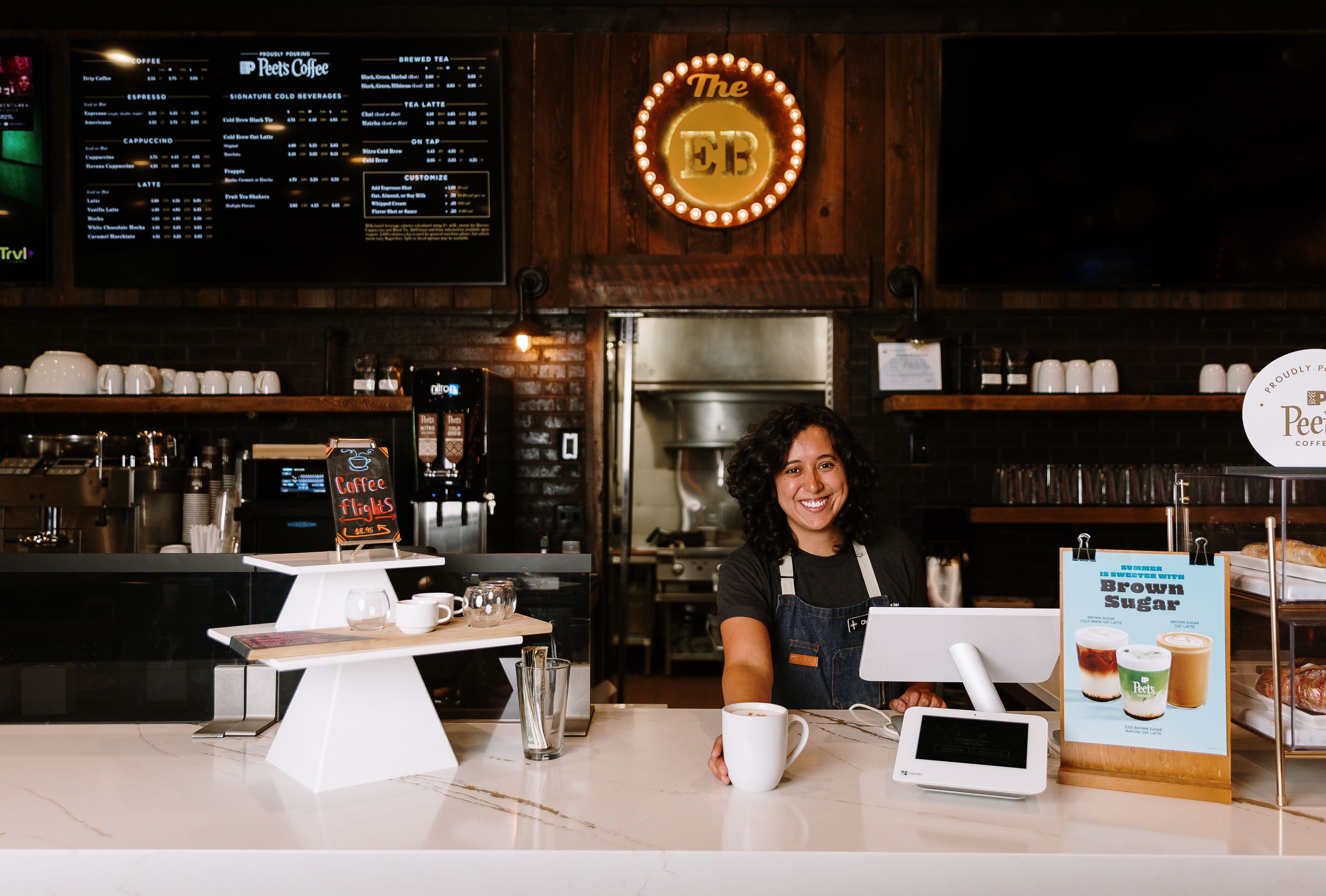 Un bancone di un caffè con una bacheca dei menu, tazze da caffè, una macchina per il caffè e un cartello con scritto 'The EB'.