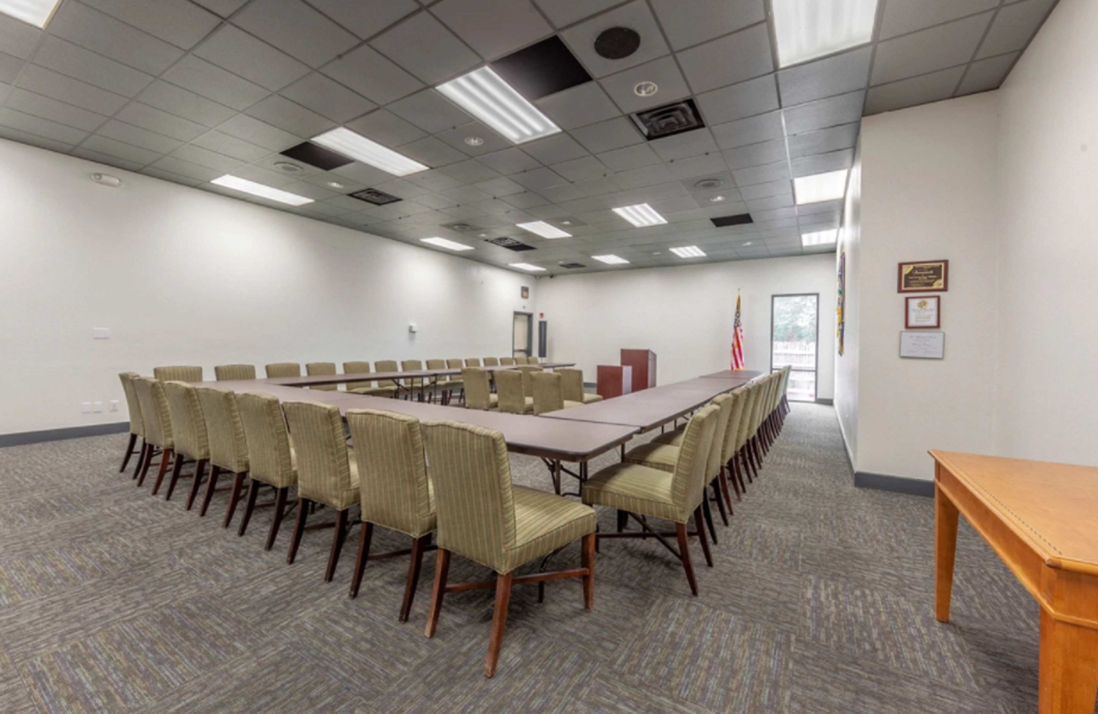 A conference room with a long table and chairs, carpeted floor, and a flag on the wall.