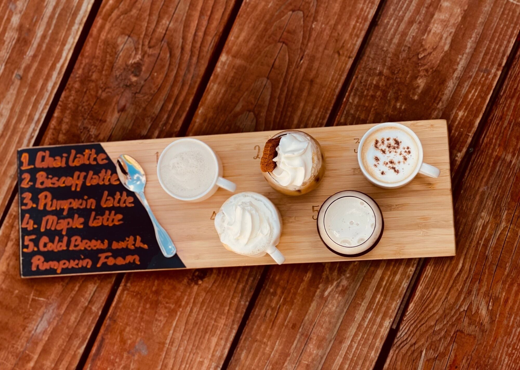 A wooden board with a list of five latte drinks and a spoon.