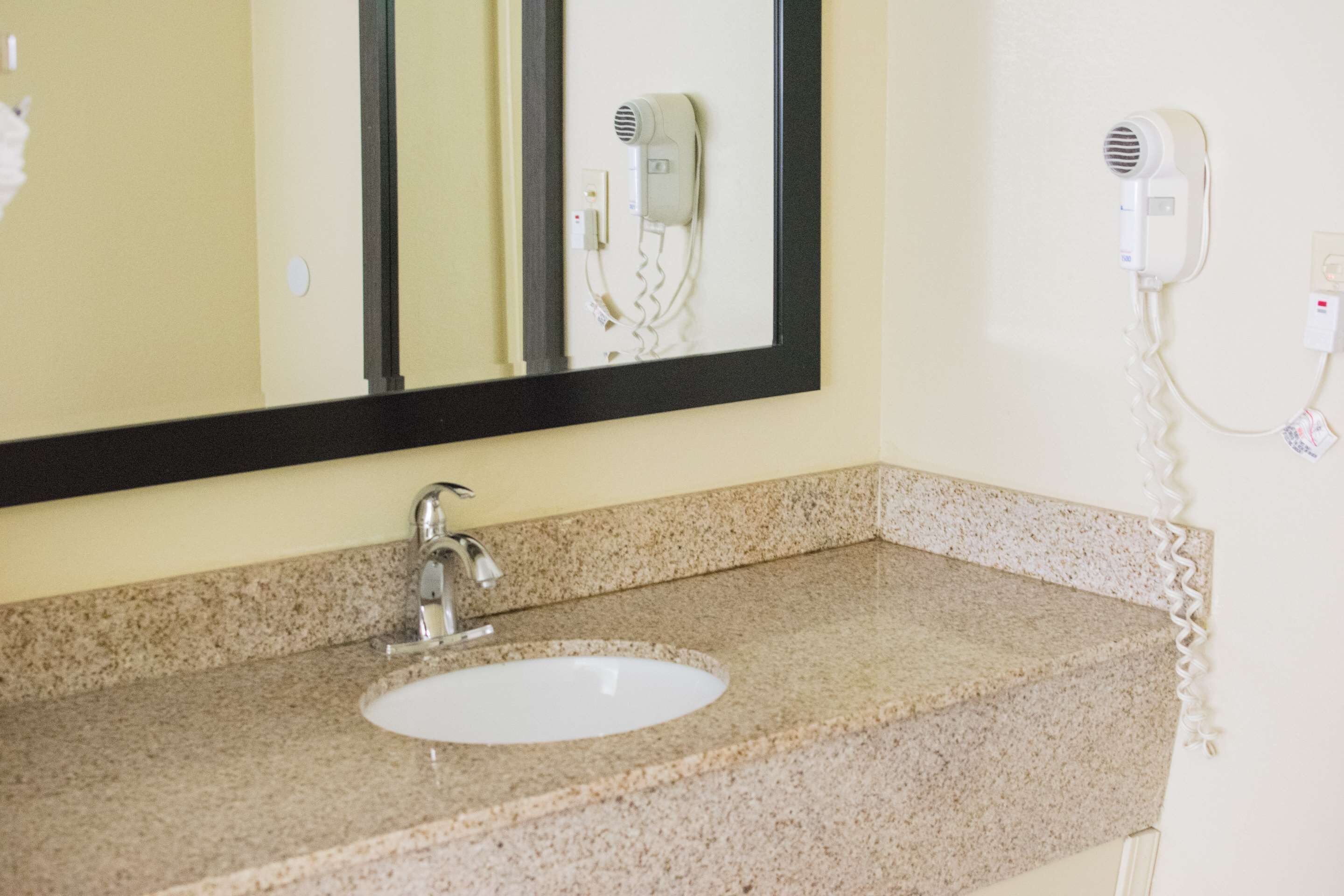 A bathroom with a granite countertop, a white sink, a mirror, and a hairdryer mounted on the wall.